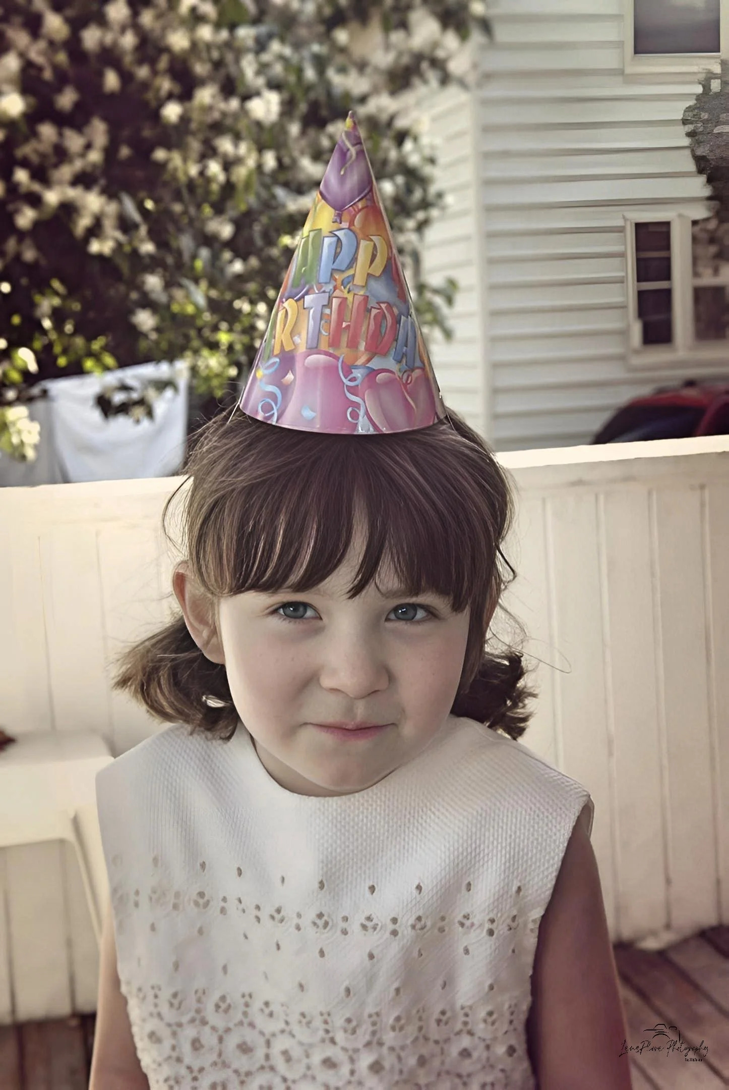 A young girl with brown hair and bangs wearing a white sleeveless dress, sitting outdoors on a deck with a white fence behind her, and a birthday hat that says 'Happy Birthday' on her head.