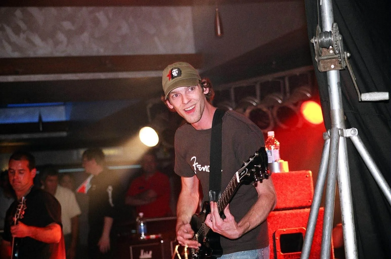 A man playing guitar on stage at a concert, smiling, wearing a baseball cap and a t-shirt, with stage lights and other musicians in the background.