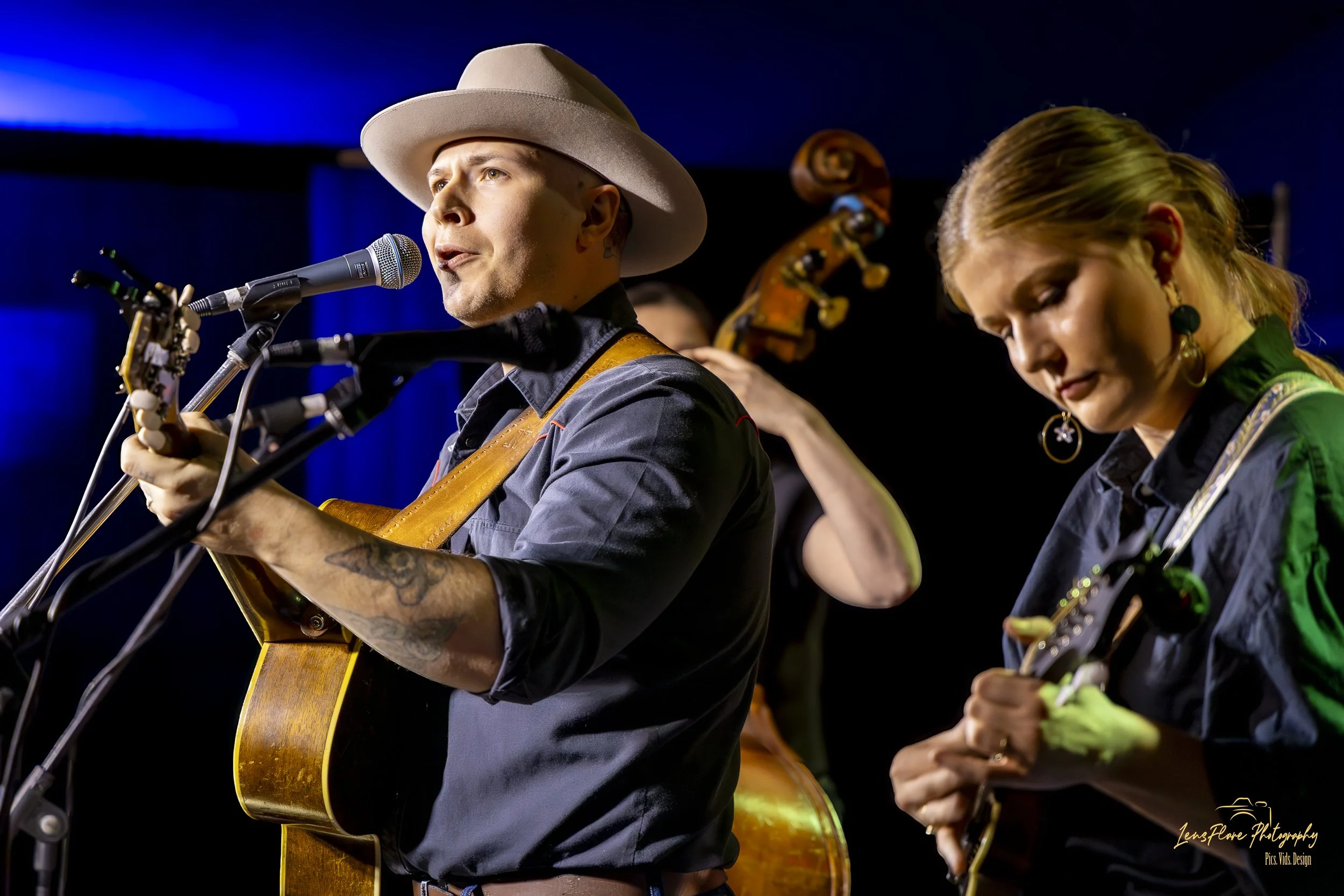 Man in a cowboy hat singing into a microphone while playing guitar, with women playing guitar beside him, on stage with blue lighting.
