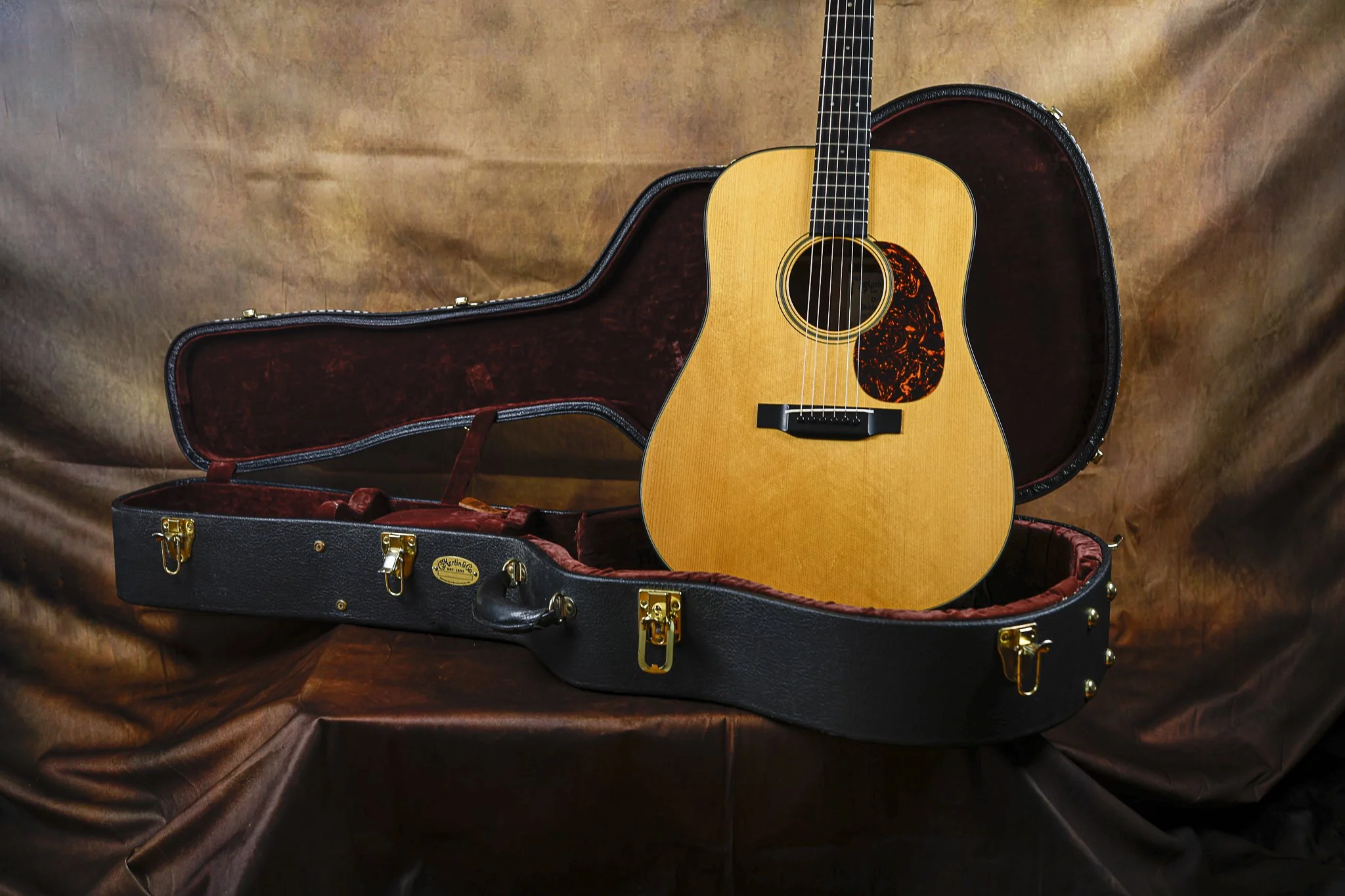 An acoustic guitar resting inside an open black case with gold hardware, set against a brown fabric background.