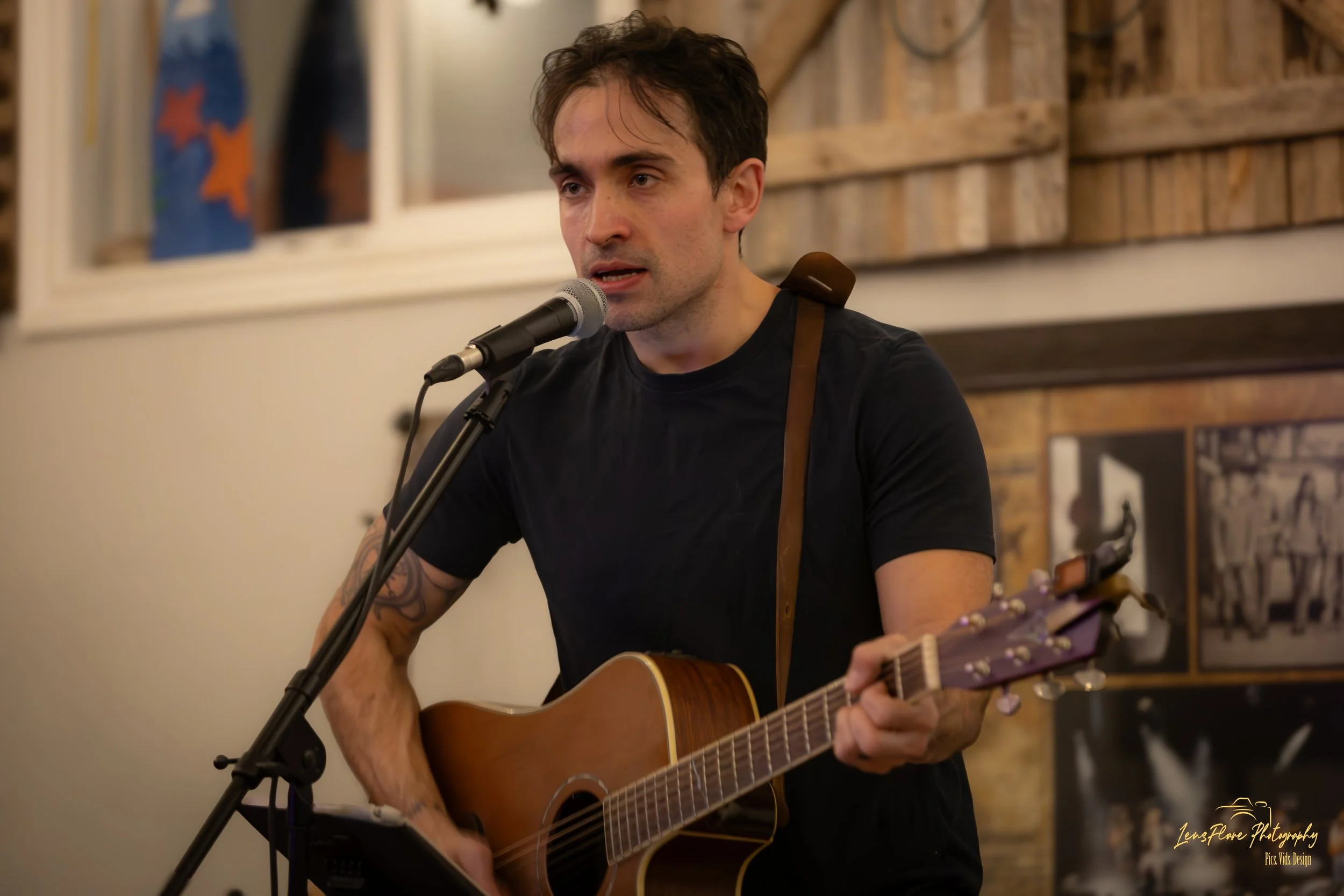 A man playing an acoustic guitar and singing into a microphone at an indoor venue with framed photos and wooden decor in the background.