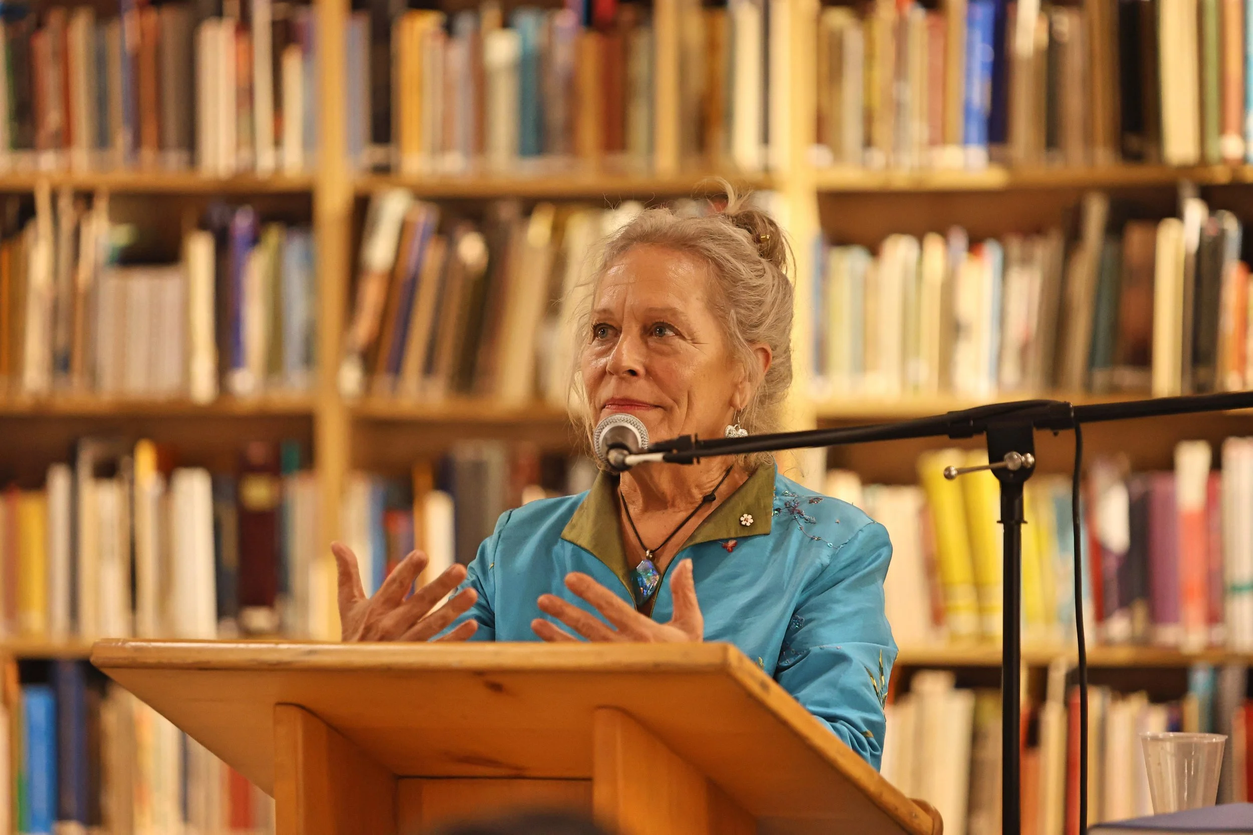 An elderly woman with gray hair tied back, wearing a turquoise jacket with embroidery, speaking into a microphone at a wooden podium in a library with shelves of books in the background.