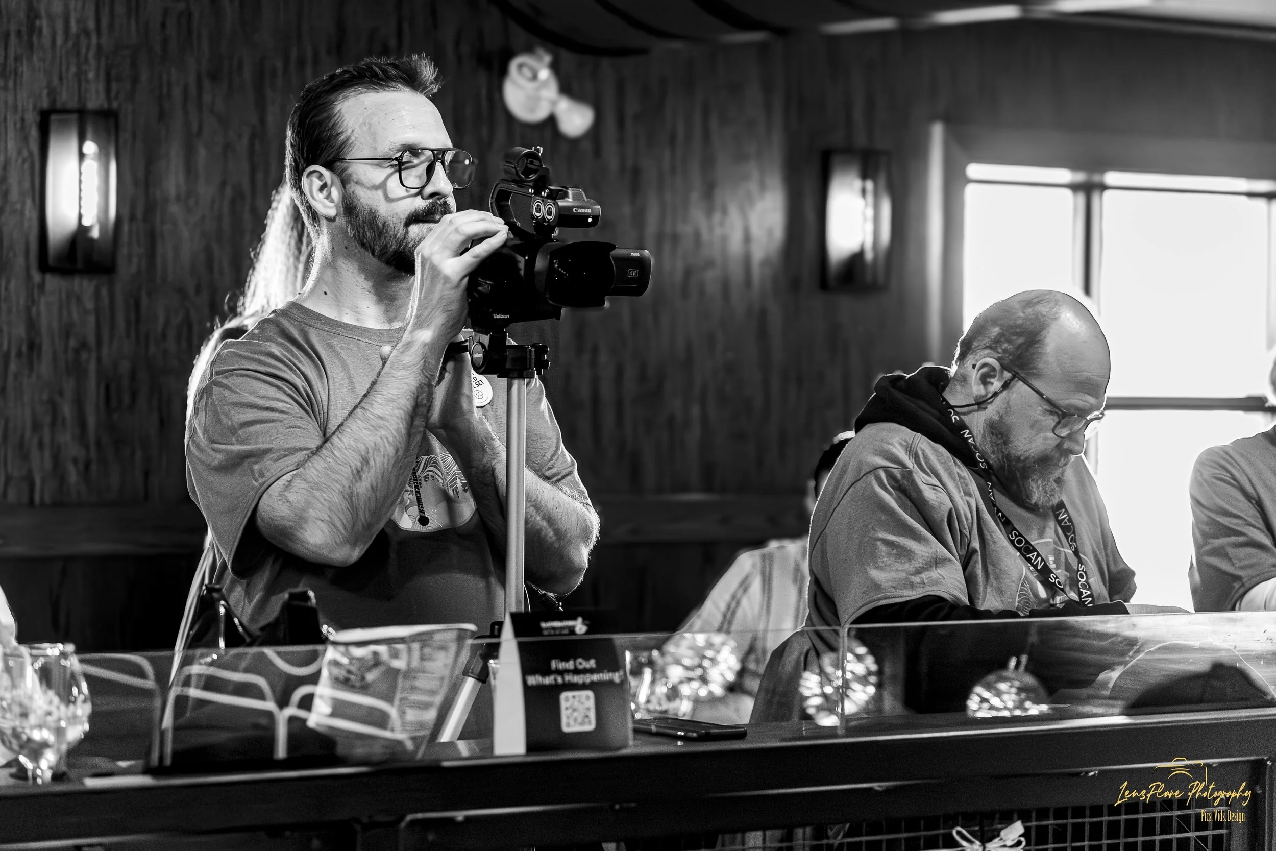 Black and white photo of a man with glasses and a beard aiming a video camera on a tripod, with people seated at a table in a room with wooden walls and large windows.