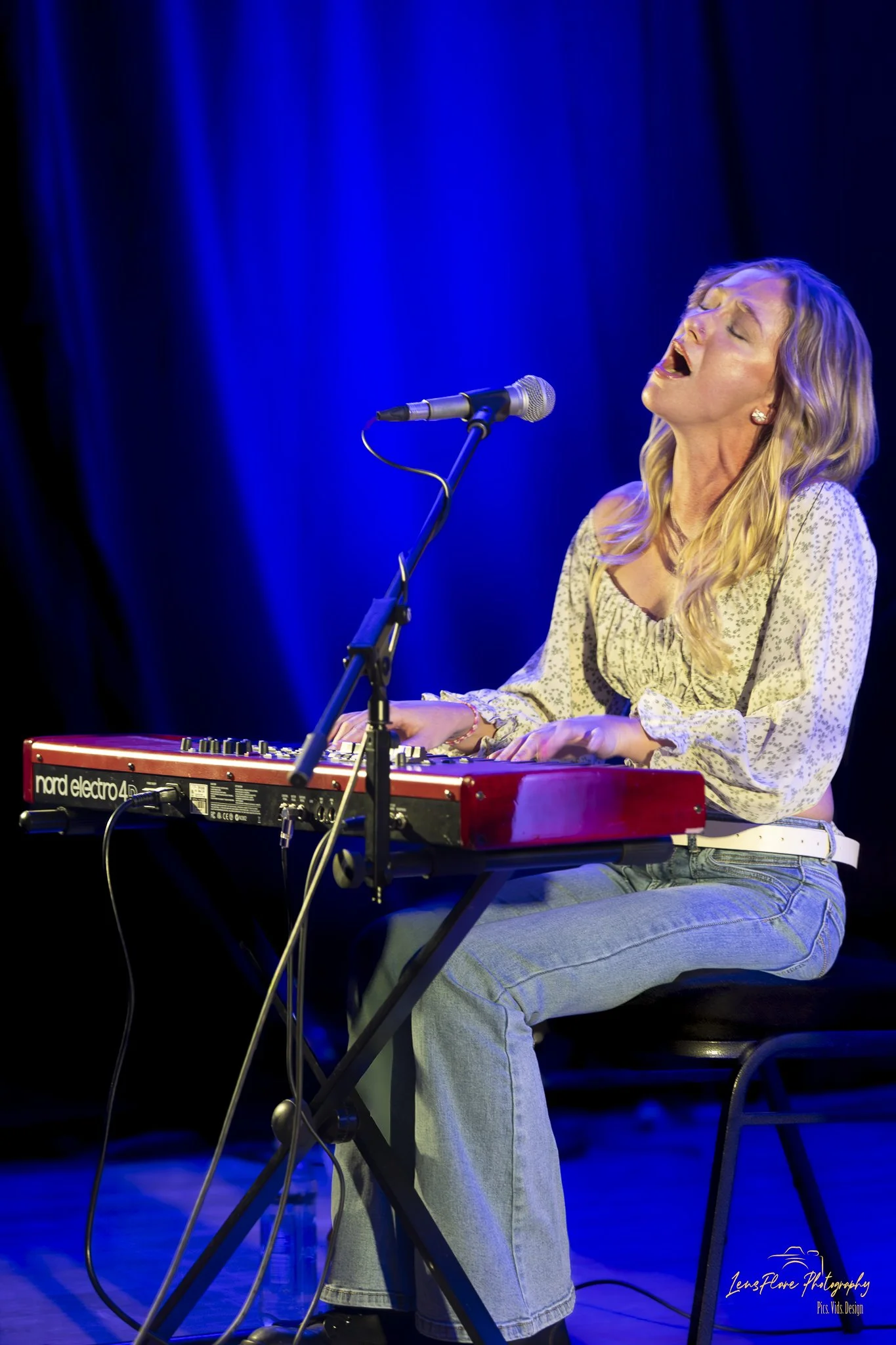 A woman singing passionately while playing a red Nord Electro 4 keyboard on a stage with blue lighting.