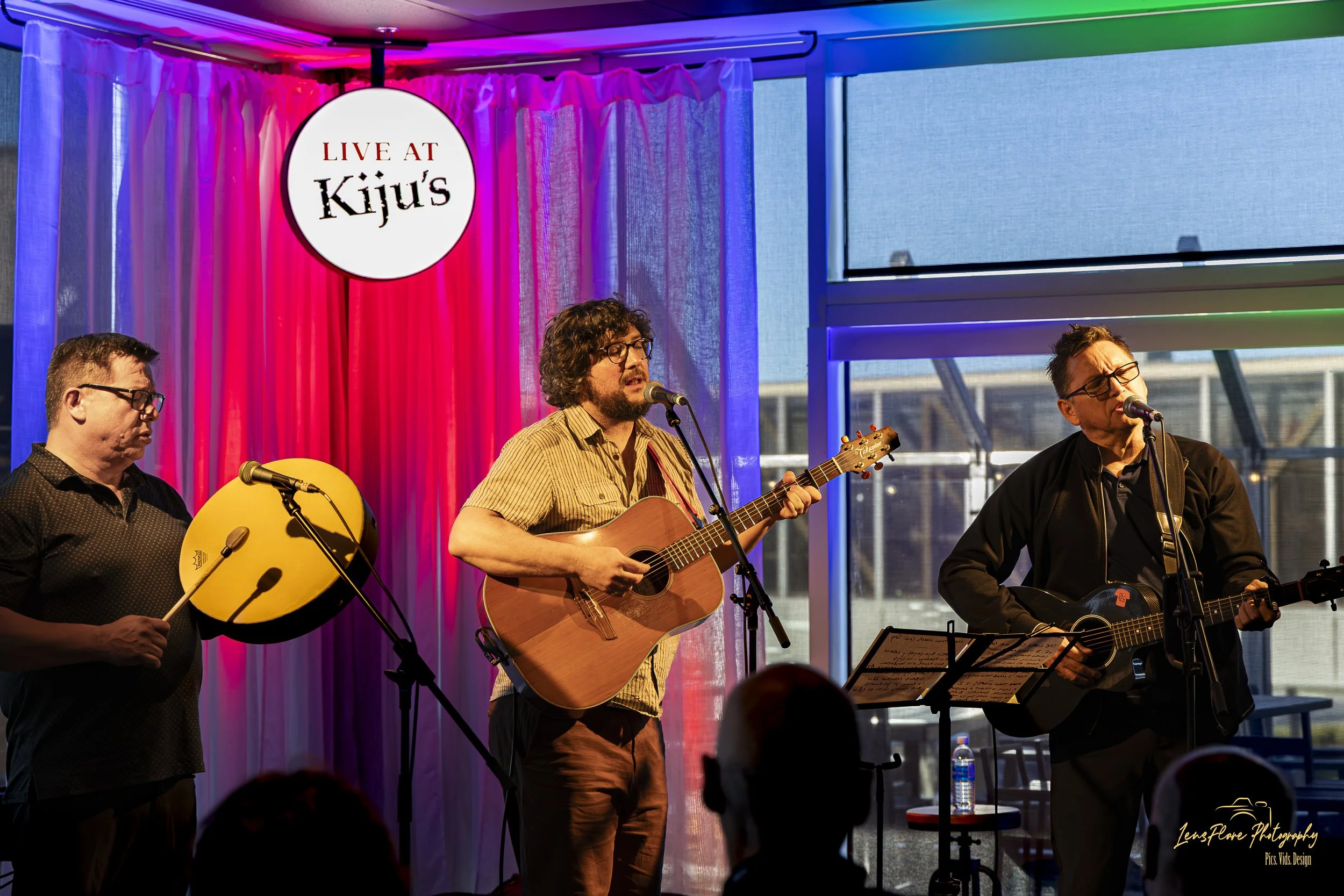 Three men are performing on stage with musical instruments, including a guitar and a tambourine. The stage has a colorful backdrop with pink, purple, and blue lighting and a circular sign that reads 'Live at Kiju's' hanging above them. Large windows 