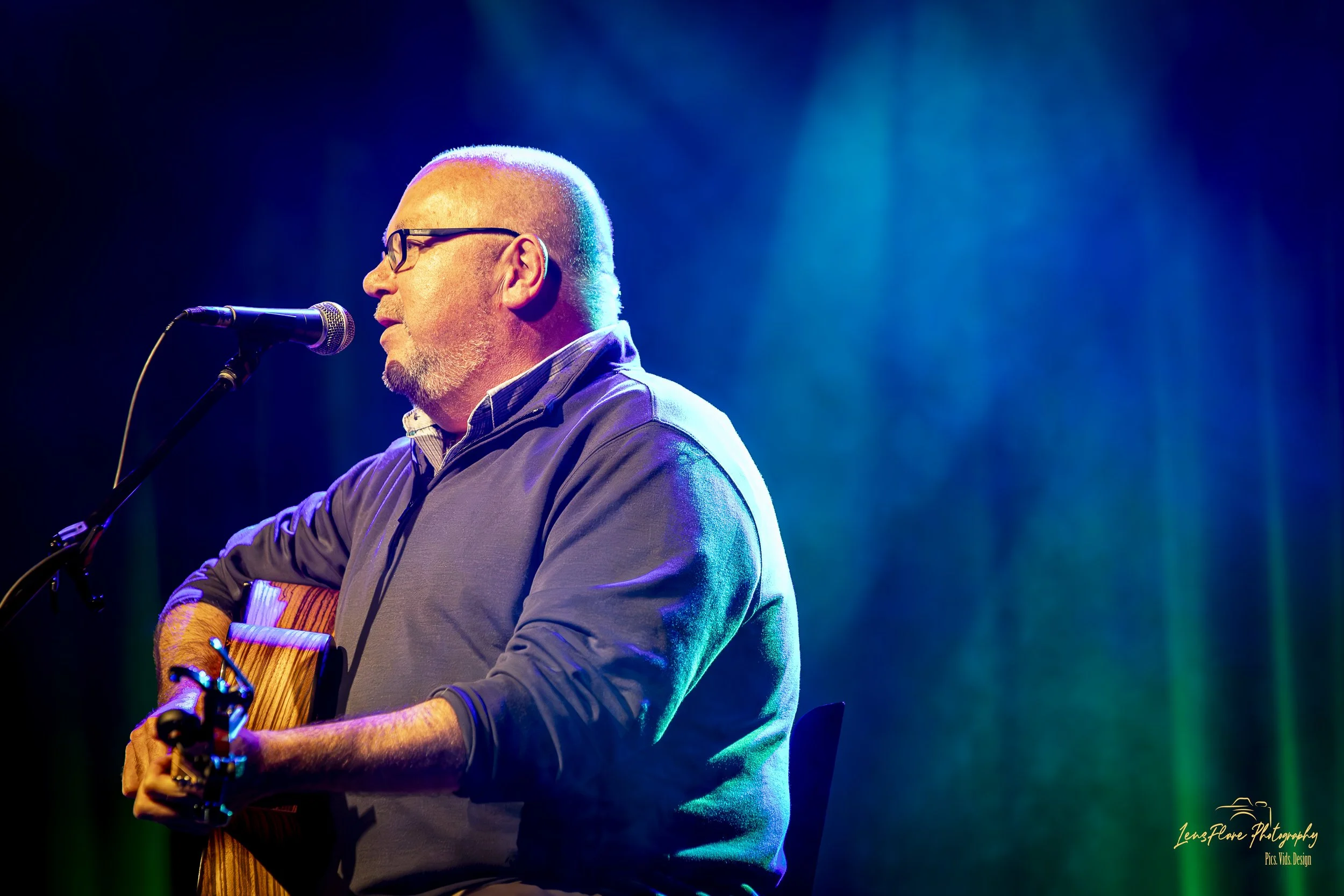 A man with glasses and a beard playing an acoustic guitar and singing into a microphone on stage, illuminated by blue and purple stage lighting.