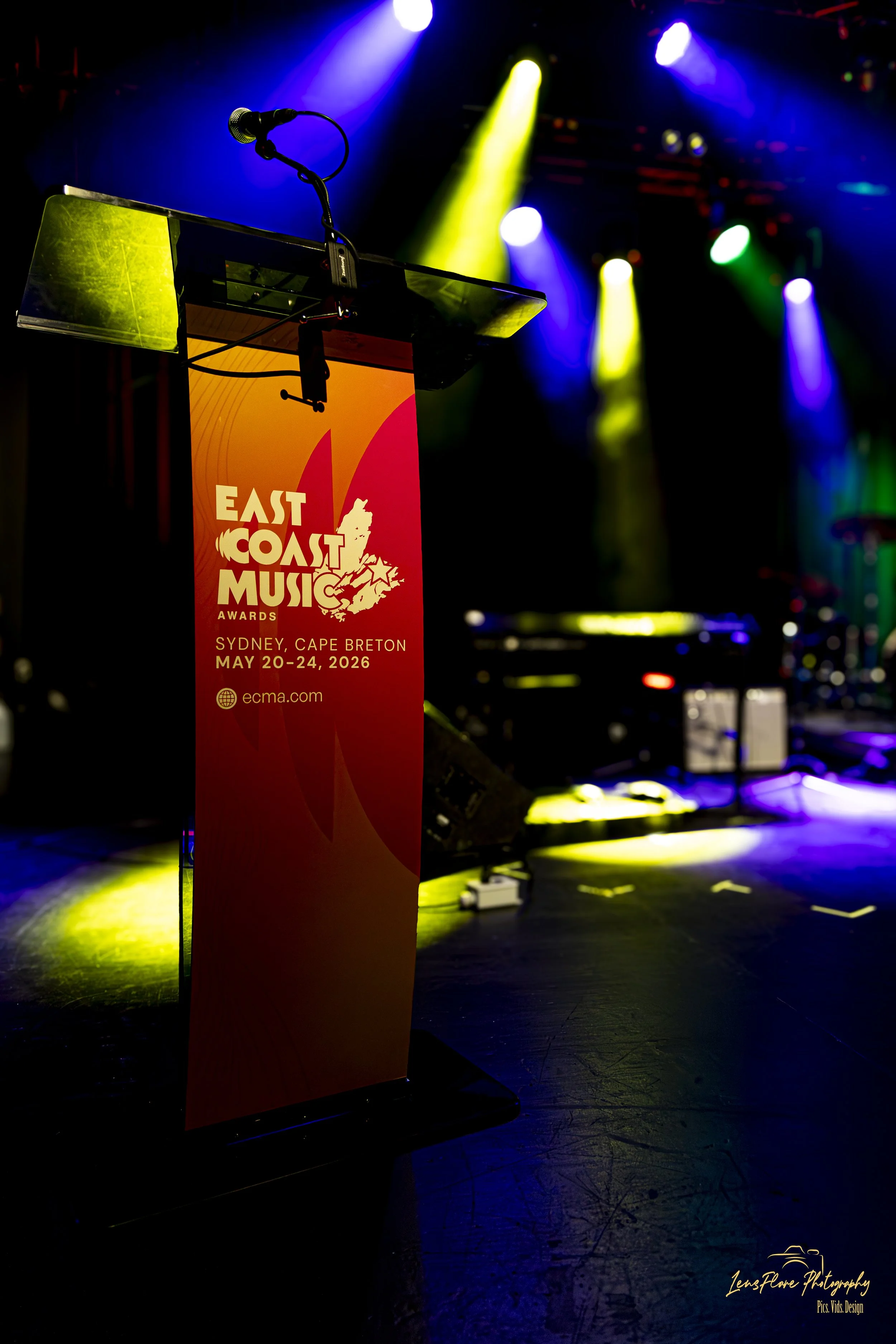A microphone on a stand at a stage with colorful stage lighting and a banner for the East Coast Music Awards in Sydney, Cape Breton, May 20-24, 2026.