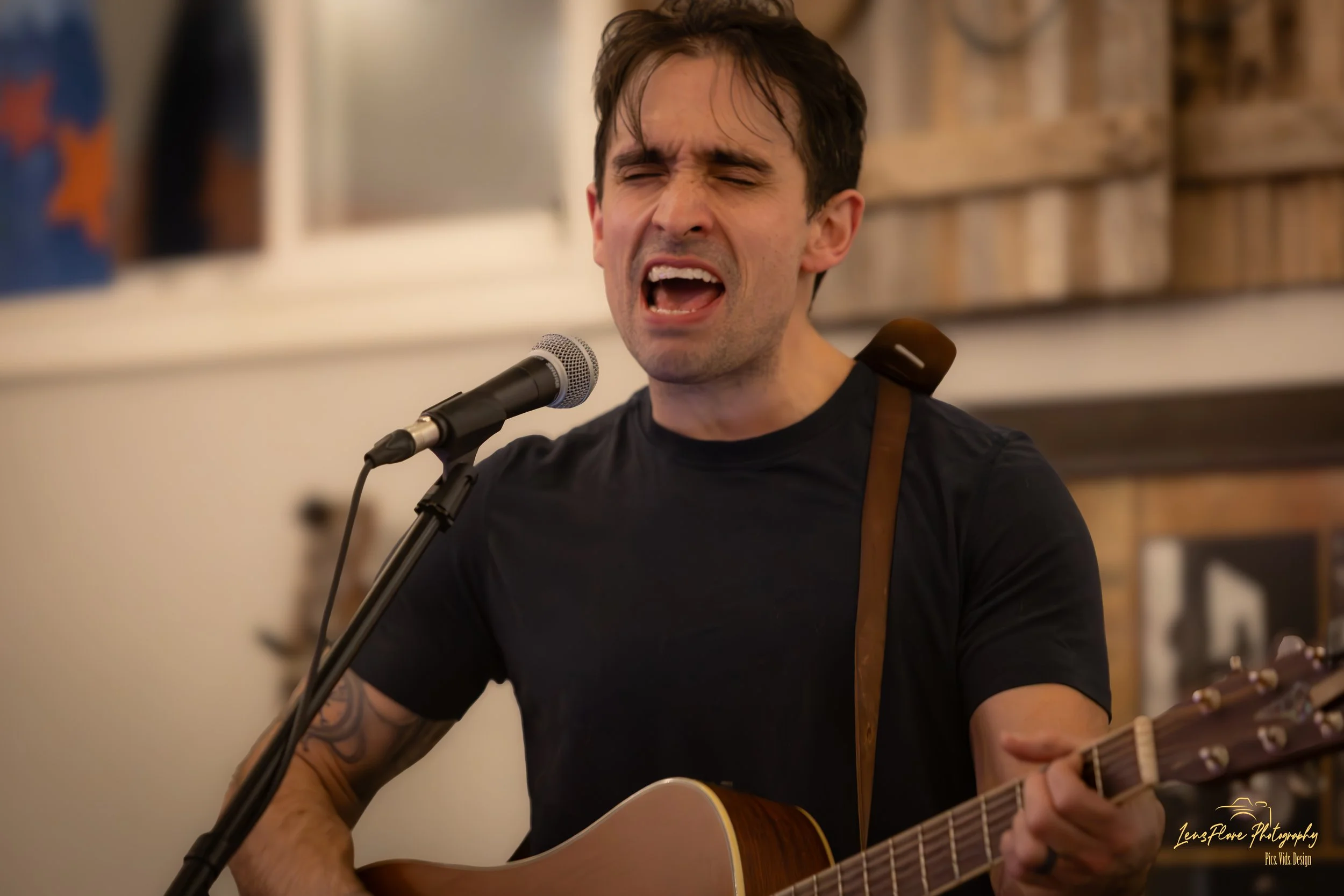 A man passionately singing into a microphone while playing an acoustic guitar, wearing a black t-shirt, with a tattoo on his right arm, in an indoor setting.