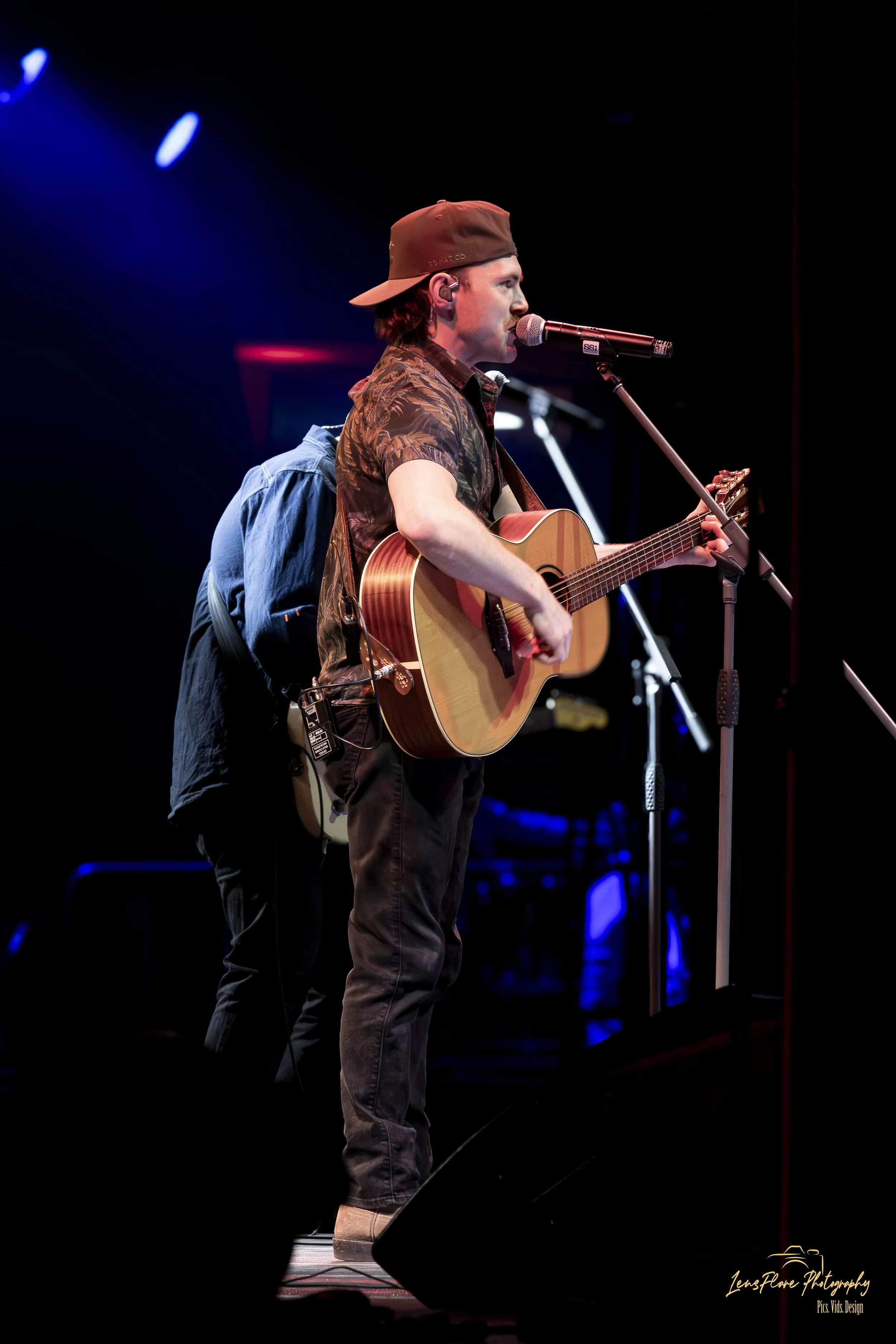 Male musician wearing a brown cap, camouflage shirt, and brown boots playing an acoustic guitar and singing into a microphone on stage with dark background and blue and red stage lights.