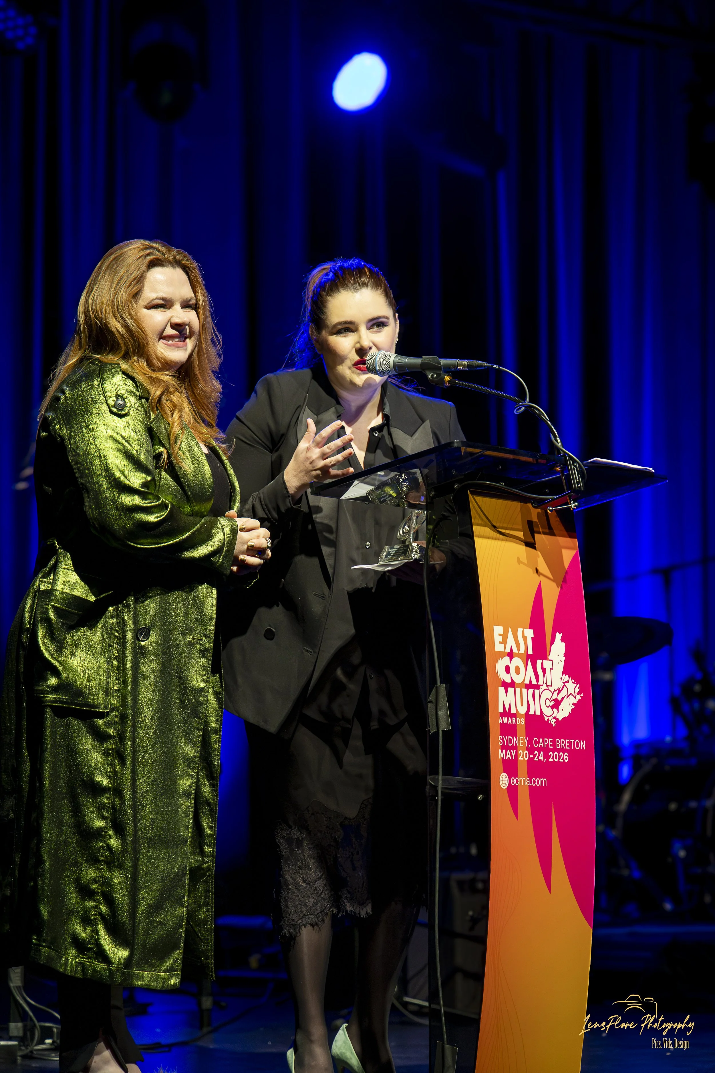 Two women standing at a podium on a stage during an award ceremony, with a Melanie headband, one in a shiny green jacket, and the other in a black jacket, speaking into a microphone. The event is the East Coast Music Awards, held in Sydney, Cape Bret