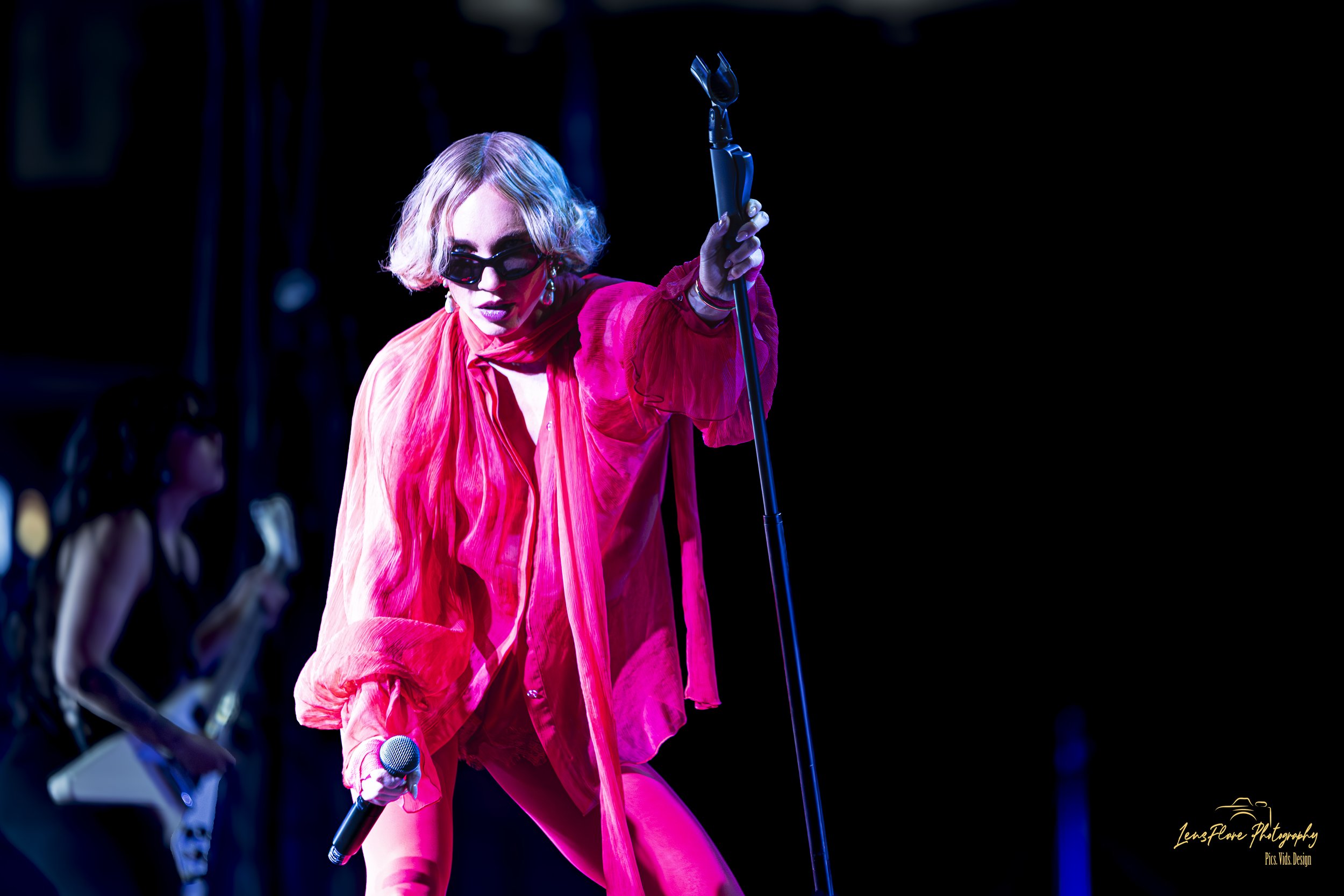 A woman wearing sunglasses and a pink outfit performing on stage with a microphone in her hand, with other performers in the background.
