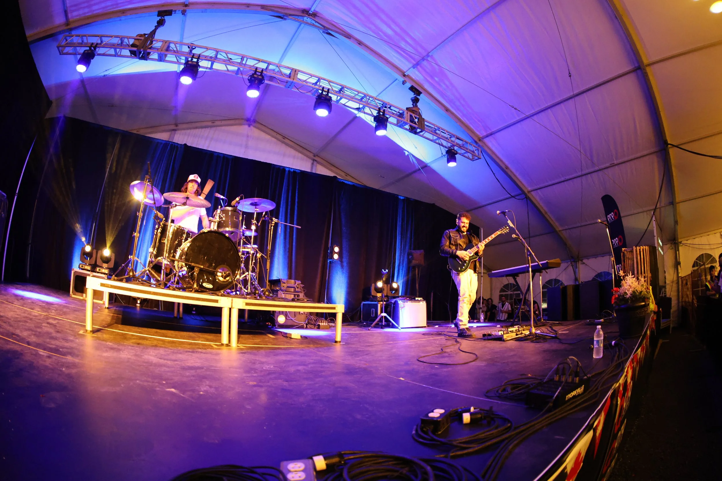 A band performing on stage inside a large tent with black curtains and colored stage lights. The drummer is on the left and the guitarist singing into a microphone on the right.