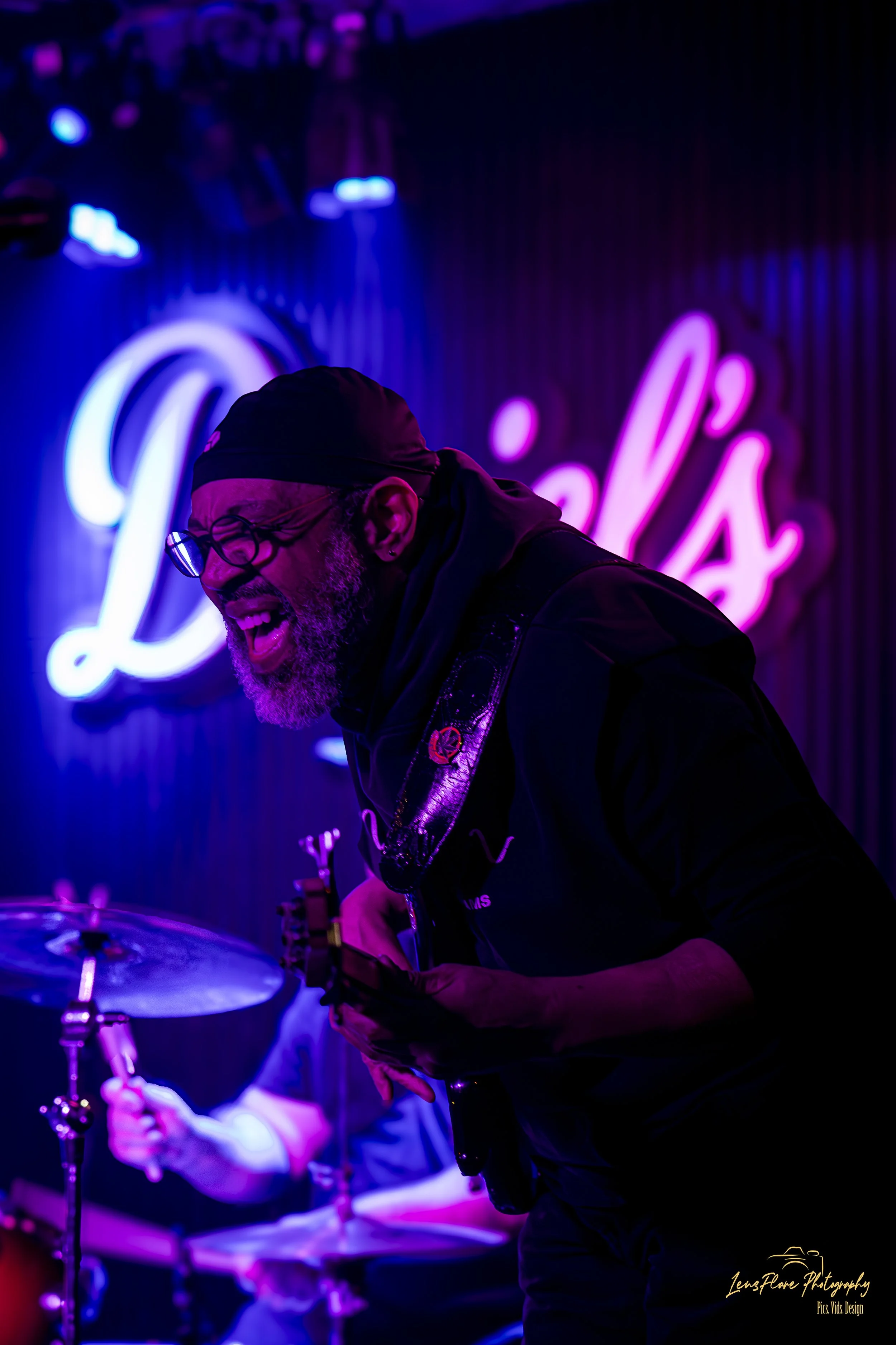 A man with glasses, a beard, and a black cap plays drums and a guitar on stage, with vibrant purple and blue neon lights in the background, and the word 'Lovel's' illuminated in pink.