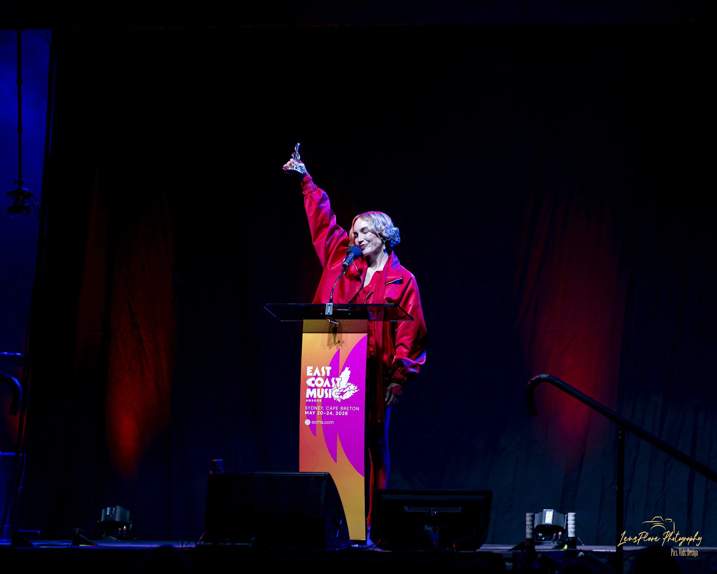 A woman in a red jacket is speaking at a podium during the East Coast Music Awards event, which takes place from May 20 to 24, 2026, in Sydney, Cape Breton. She is holding an award in her raised right hand and smiling.