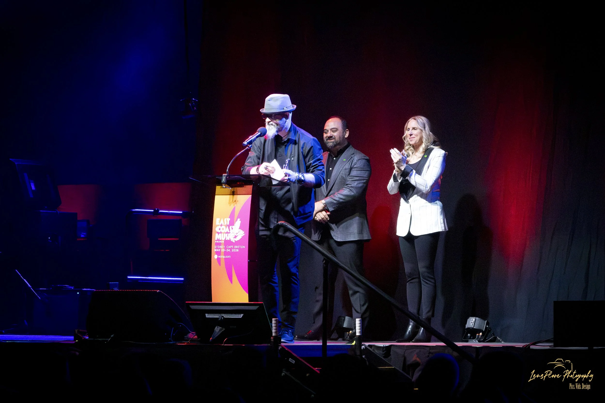 A man with a beard and sunglasses, wearing a hat, stands at a podium reading from a paper, flanked by a man and a woman on a stage with a dark red curtain backdrop. The woman claps and both men appear to be smiling. A sign on the podium indicates an 