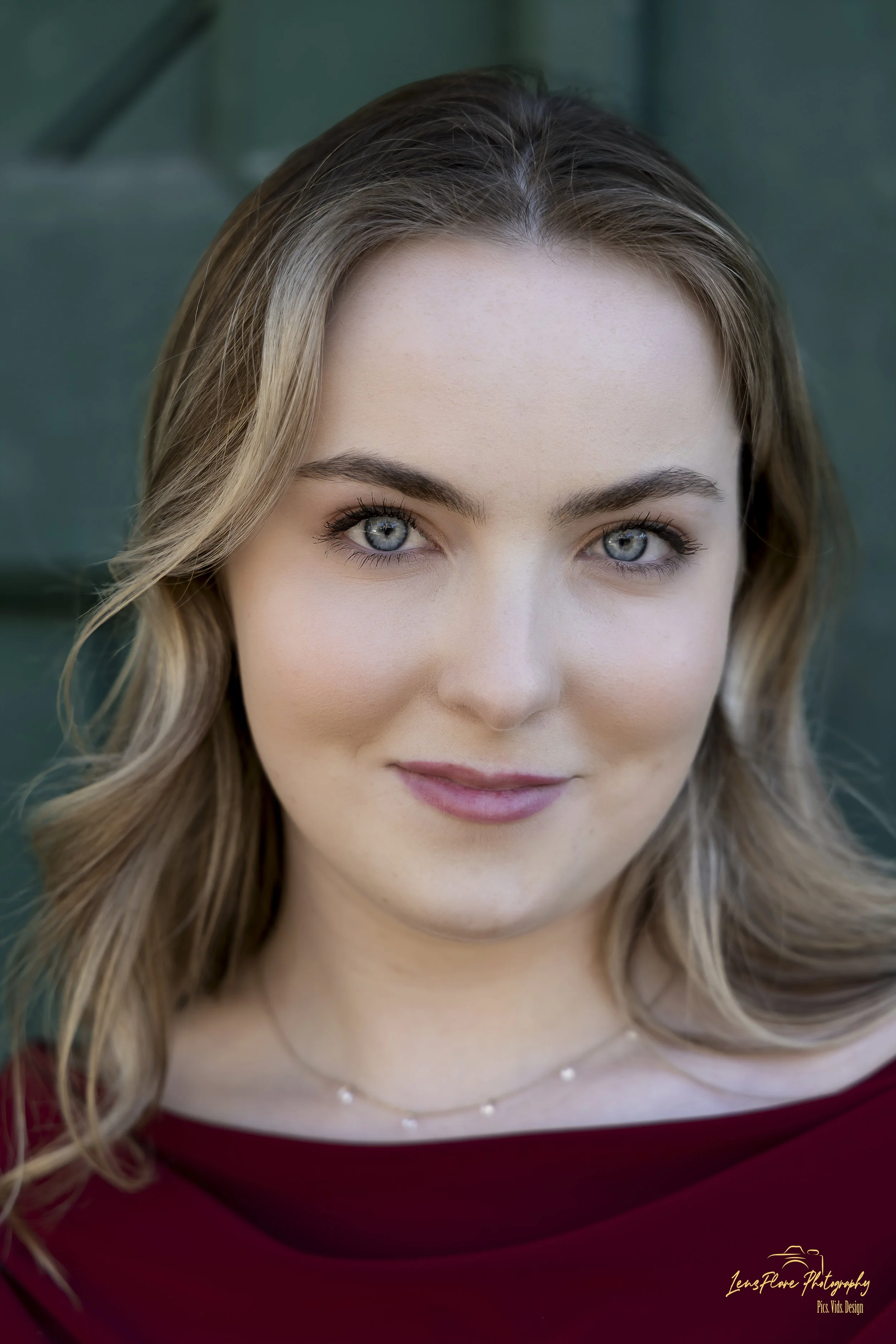 Close-up of a young woman with blue eyes and wavy, blonde hair in front of a green background.