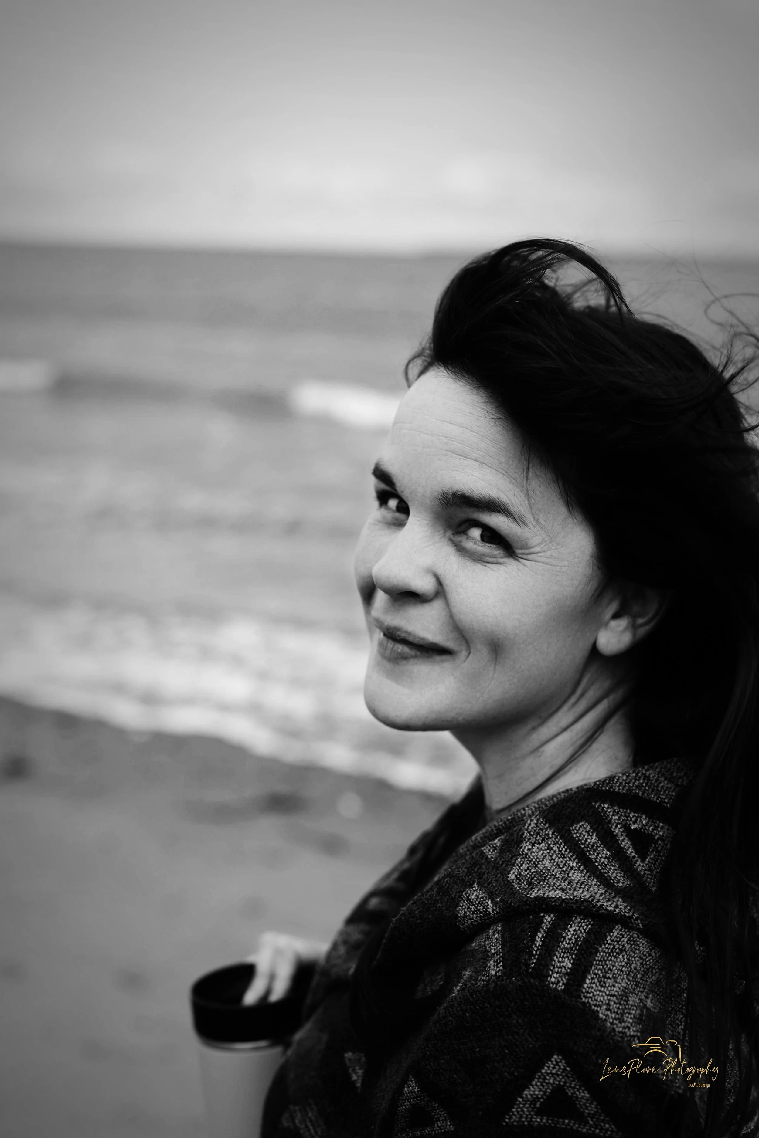 Black and white portrait of a woman with long hair outdoors at the beach, looking over her shoulder with a slight smile.