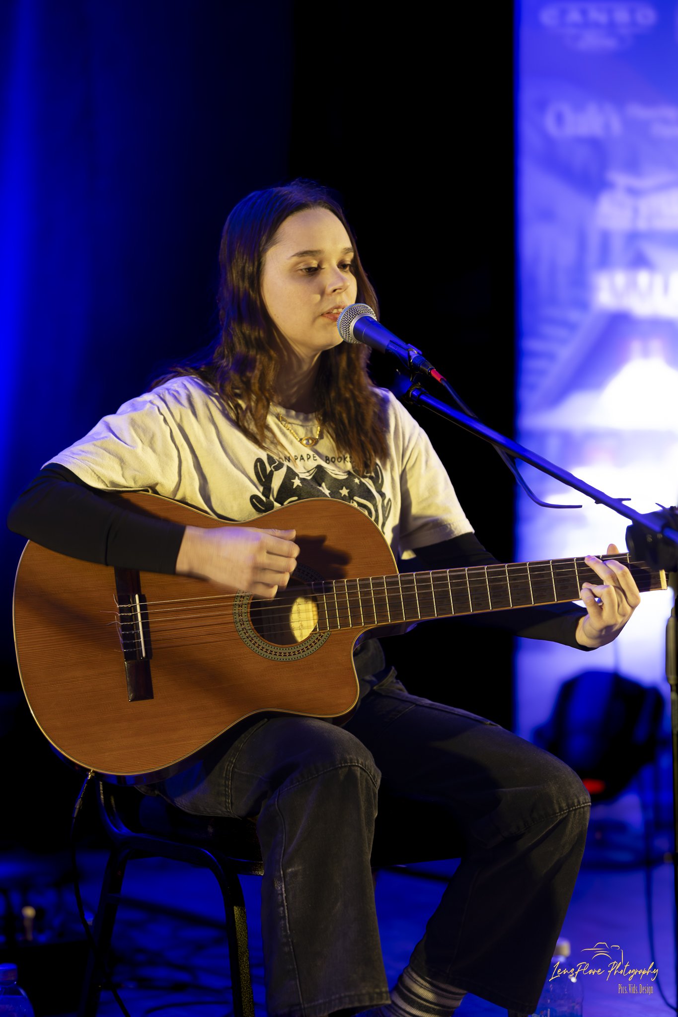 A young woman with long wavy hair sitting on a chair, playing an acoustic guitar and singing into a microphone on stage with blue lighting.