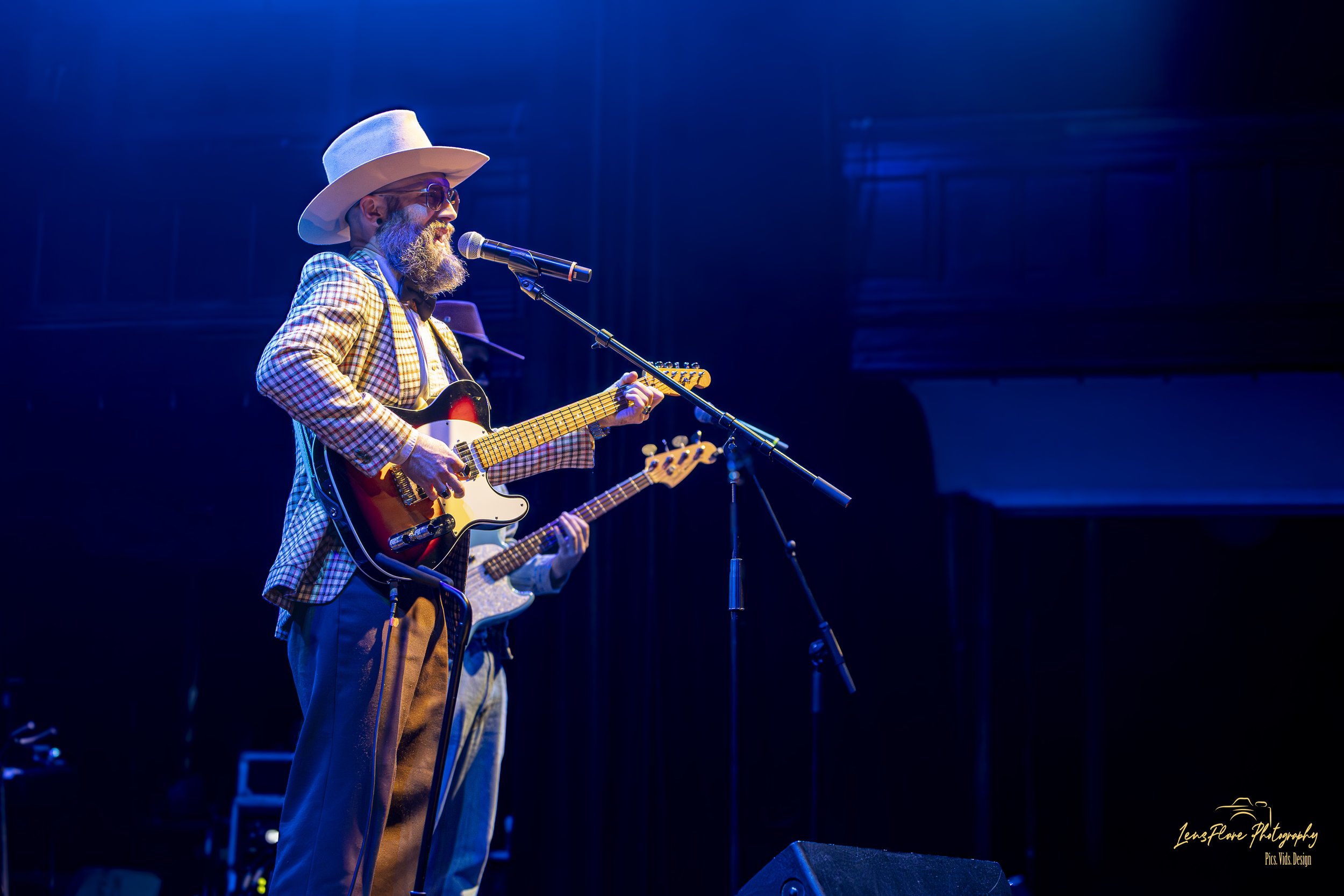 A musician wearing a large white hat, sunglasses, and a checkered blazer plays an electric guitar on stage under blue lighting.