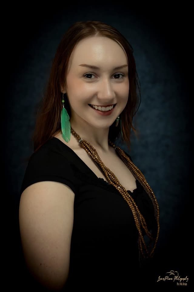 Portrait of a young woman with red hair, wearing a black top, green feather earrings, and layered beaded necklaces, smiling against a dark background.