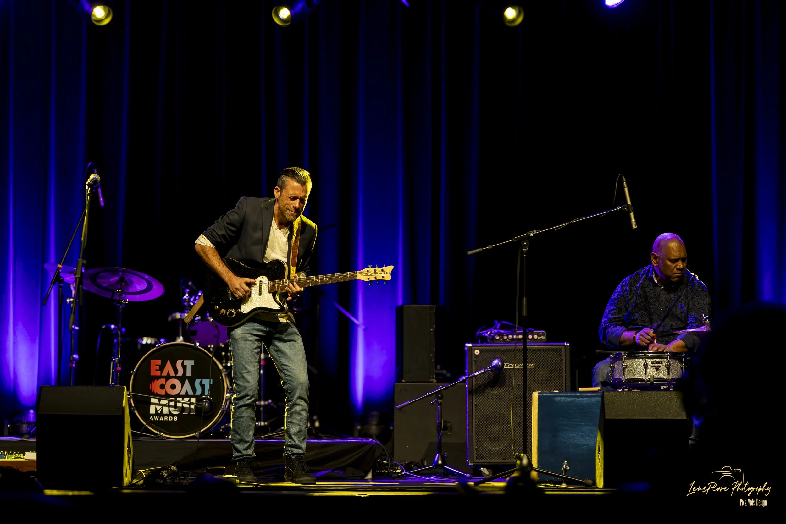 Two musicians performing on a stage with blue curtains. The musician on the left is playing an electric guitar and appears to be singing, wearing a black jacket and jeans. The musician on the right is sitting and playing drums, wearing a dark pattern