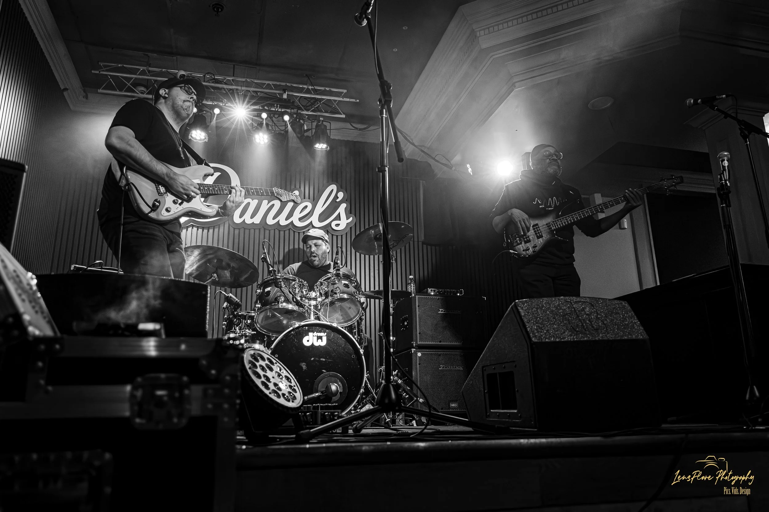A black and white photo of a live band performing on stage, including a guitarist, bassist, and drummer, with stage lights and a sign that reads 'Daniel's' in the background.