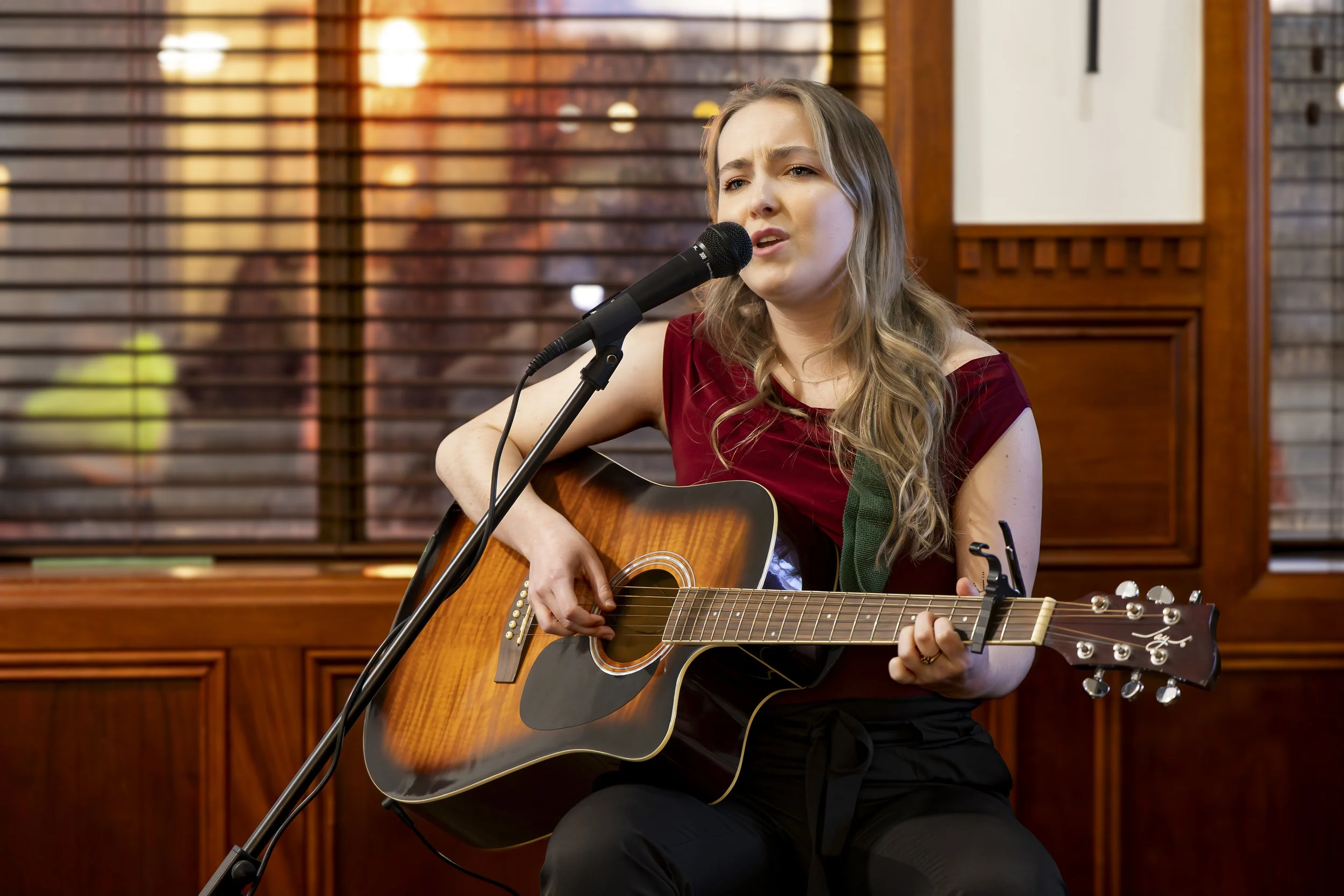A young woman with long blonde hair singing into a microphone while playing an acoustic guitar. She is wearing a red top and is seated indoors against a wooden background with blinds and warm lighting.