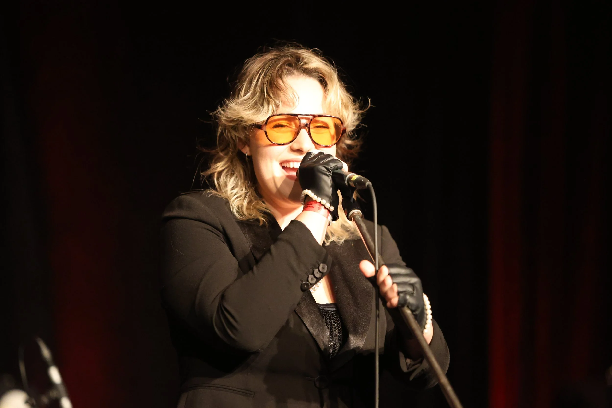 Woman singing into a microphone, wearing orange sunglasses, black blazer, gloves, and pearl bracelet, on a stage with a dark background.