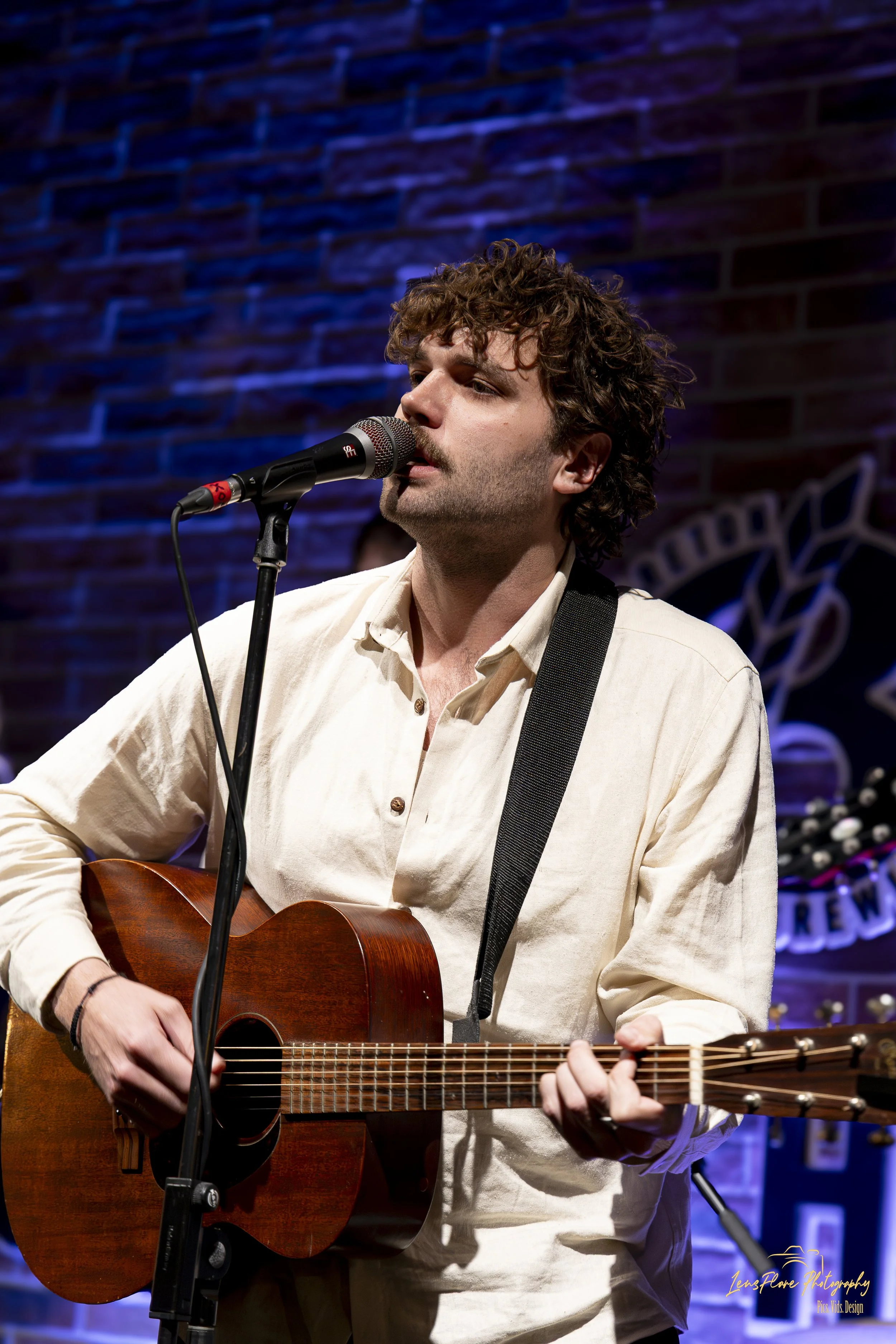 Young man with curly hair playing an acoustic guitar and singing into a microphone on stage with a brick wall background.