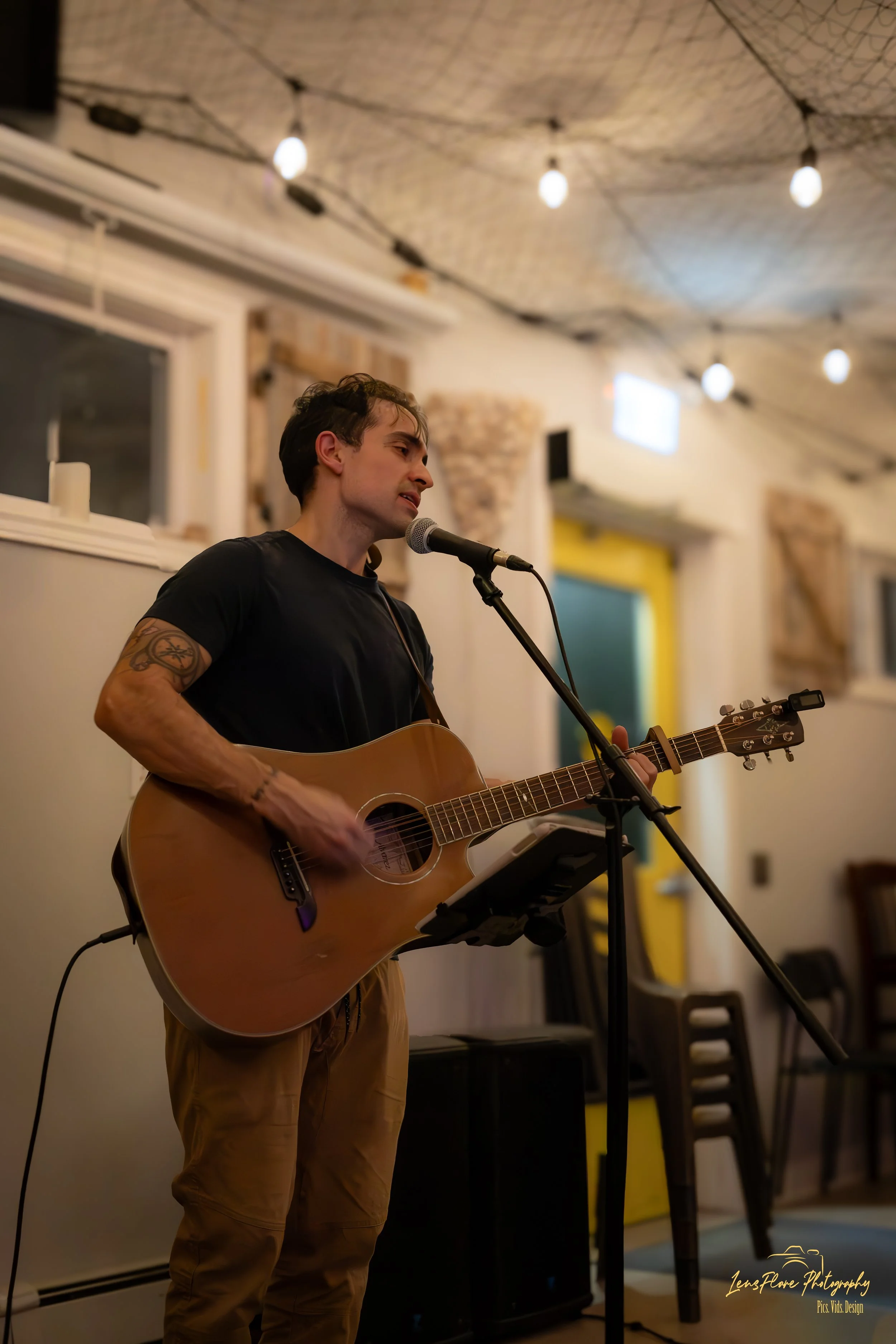 A man with tattoos on his right arm is singing and playing an acoustic guitar at a microphone in a cozy, dimly lit indoor space with string lights on the ceiling.