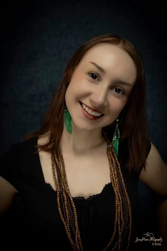 Portrait of a woman with shoulder-length reddish-brown hair, smiling, wearing green feather earrings and multiple beaded necklaces, against a dark background.