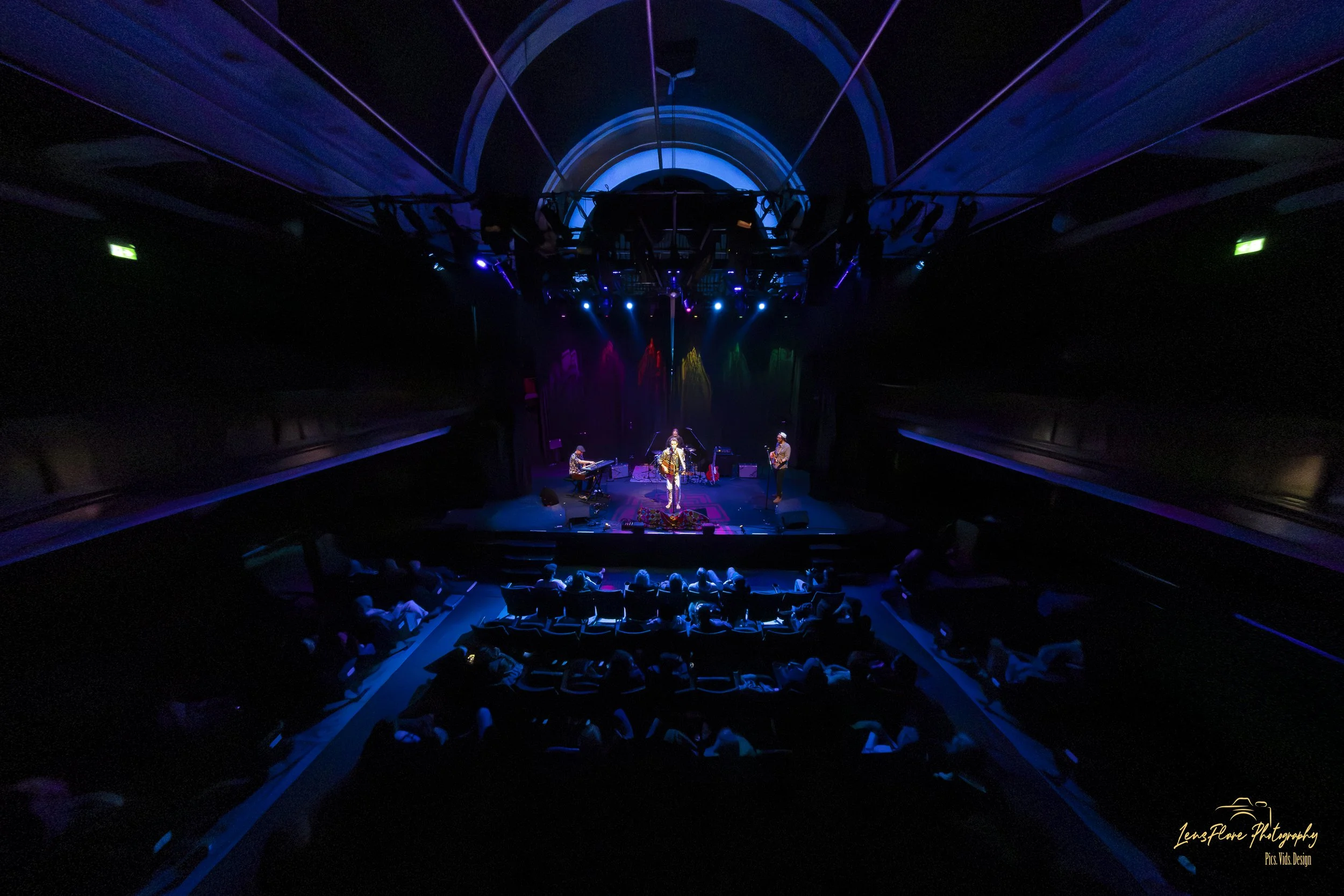 A live music performance on stage with band members playing instruments, illuminated by colorful stage lights, in a dark theater with an audience seated in front.