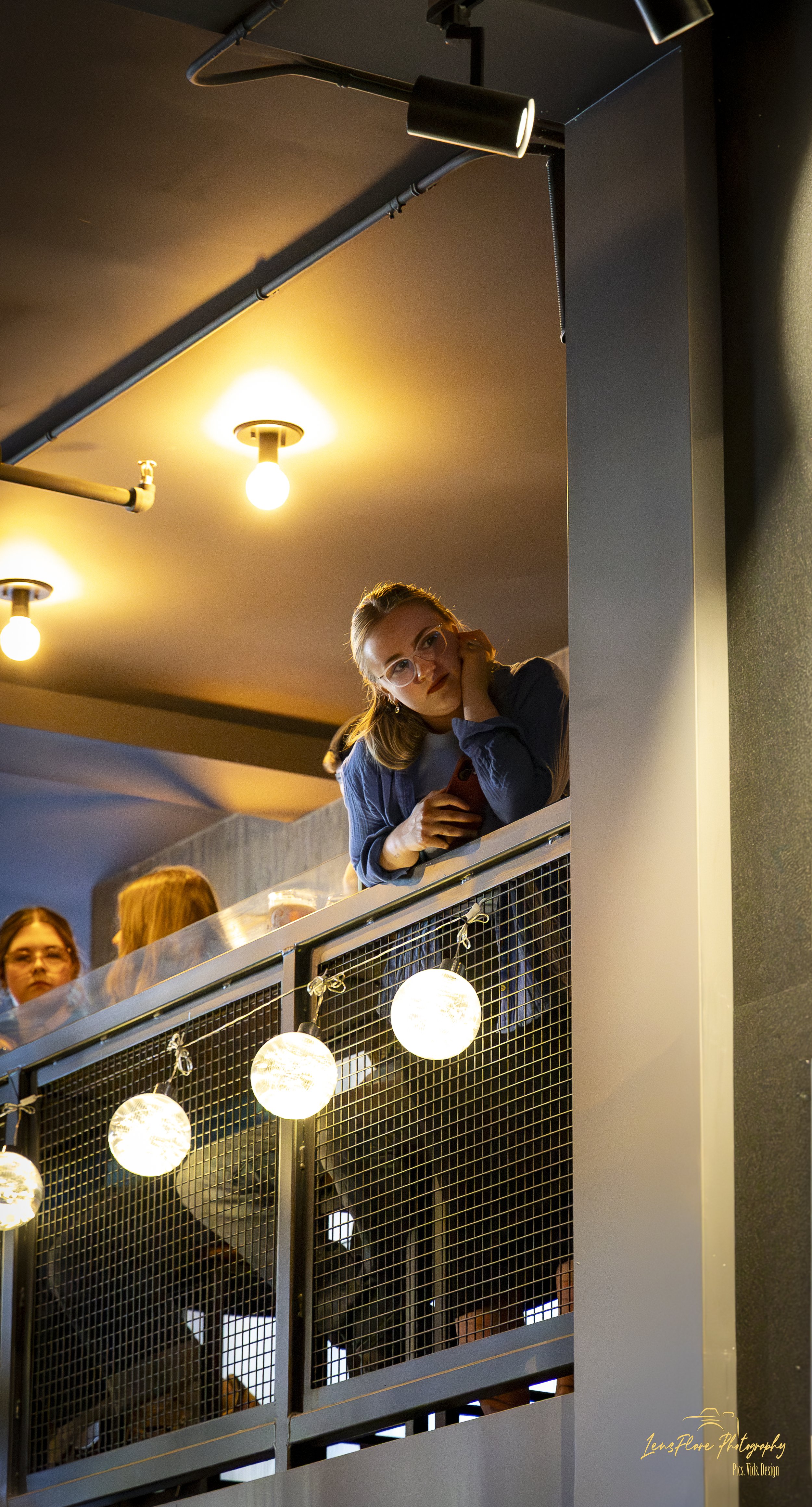 A young woman with glasses leaning on a balcony railing, holding a phone, looking at the camera, with warm lights hanging on the balcony.