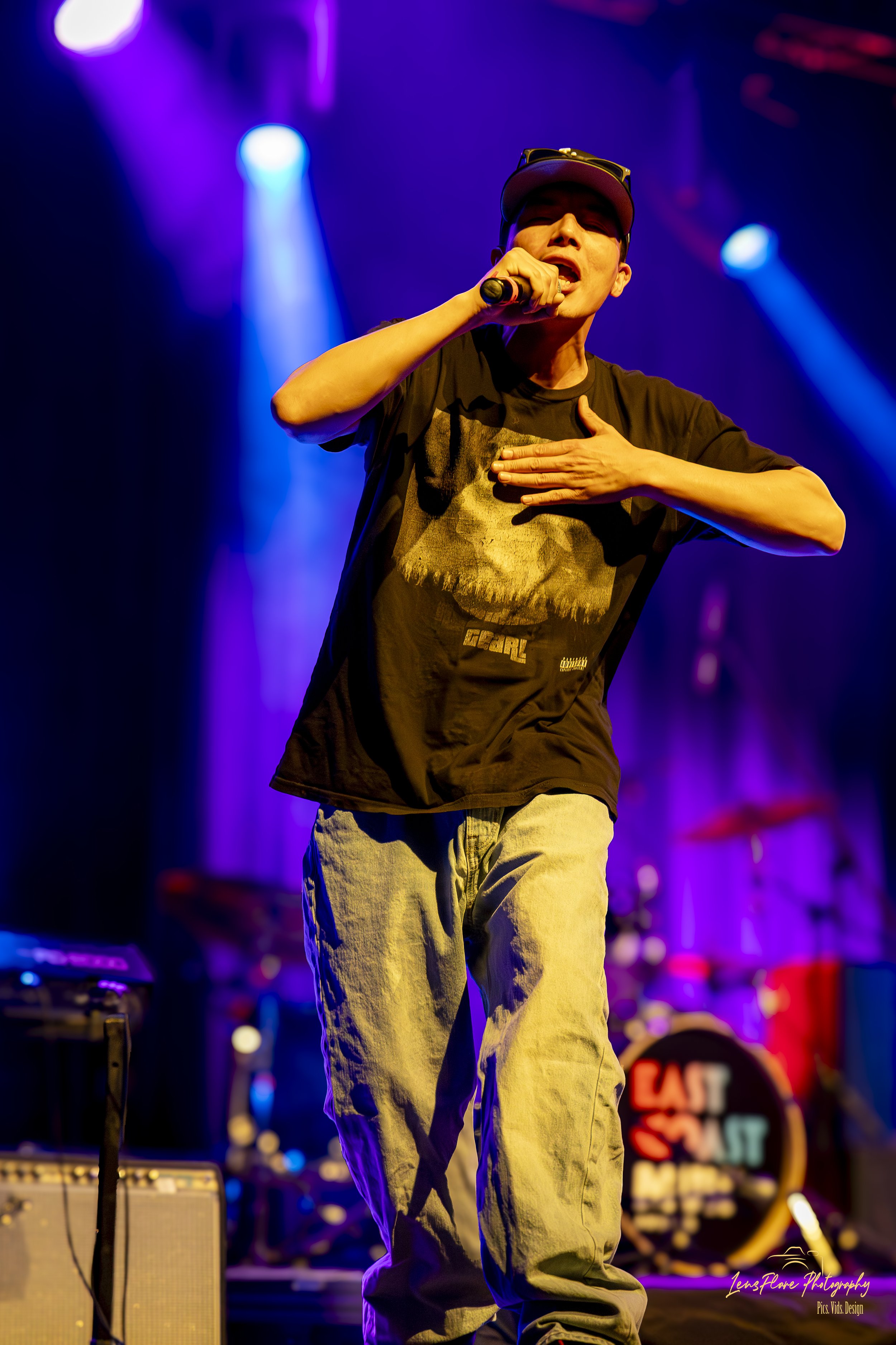 Male performer singing into microphone on stage with colorful purple and blue lighting, wearing a baseball cap, black T-shirt, and beige pants.