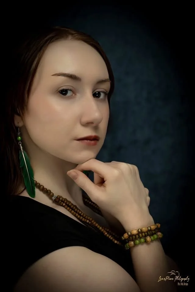 Portrait of a young woman with brown hair and light skin, wearing green earrings, a beaded necklace, and bracelets, with a thoughtful expression against a dark background.