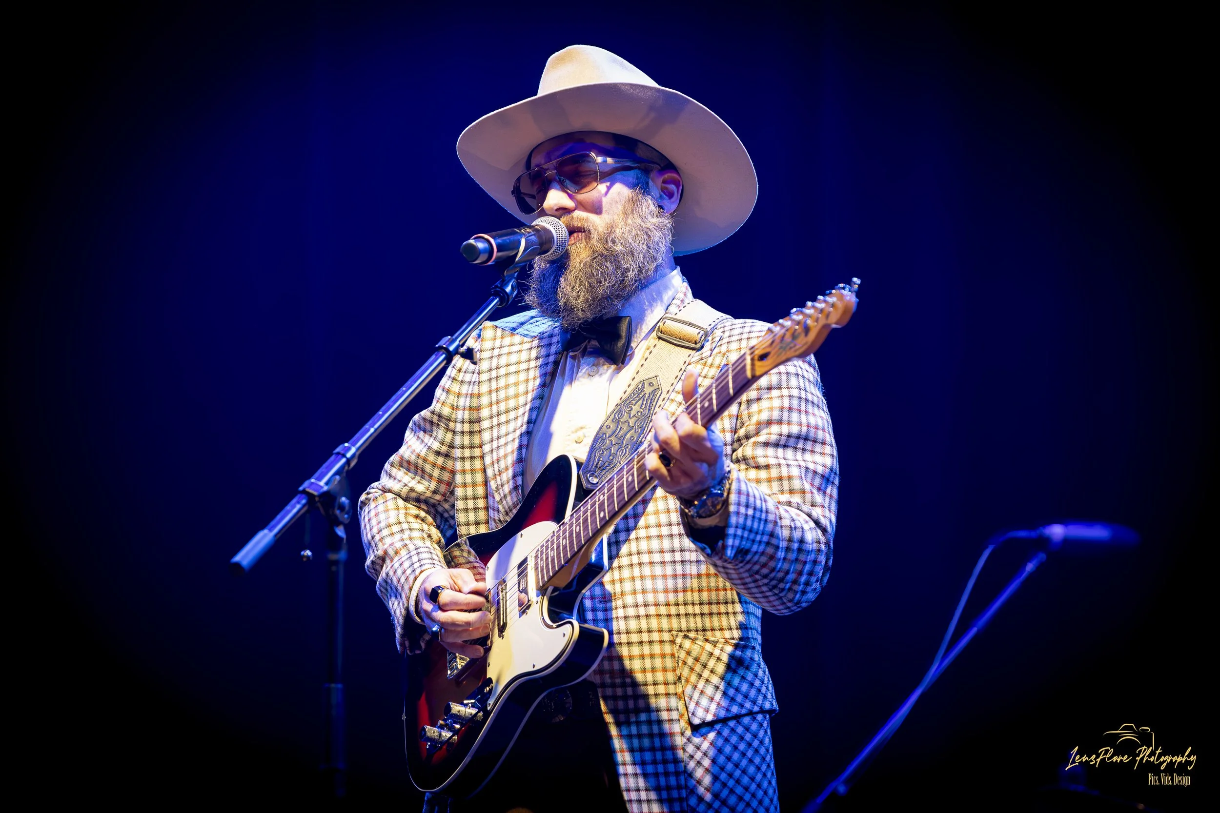 A man performing on stage, wearing a plaid suit, white cowboy hat, sunglasses, and a beard, playing an electric guitar and singing into a microphone against a dark background.