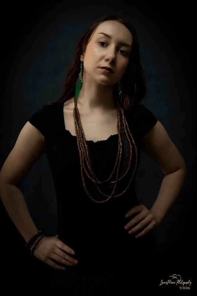 Portrait of a woman with long brown hair, wearing a black top, layered necklaces, and earrings, posing with hands on hips against a dark background.