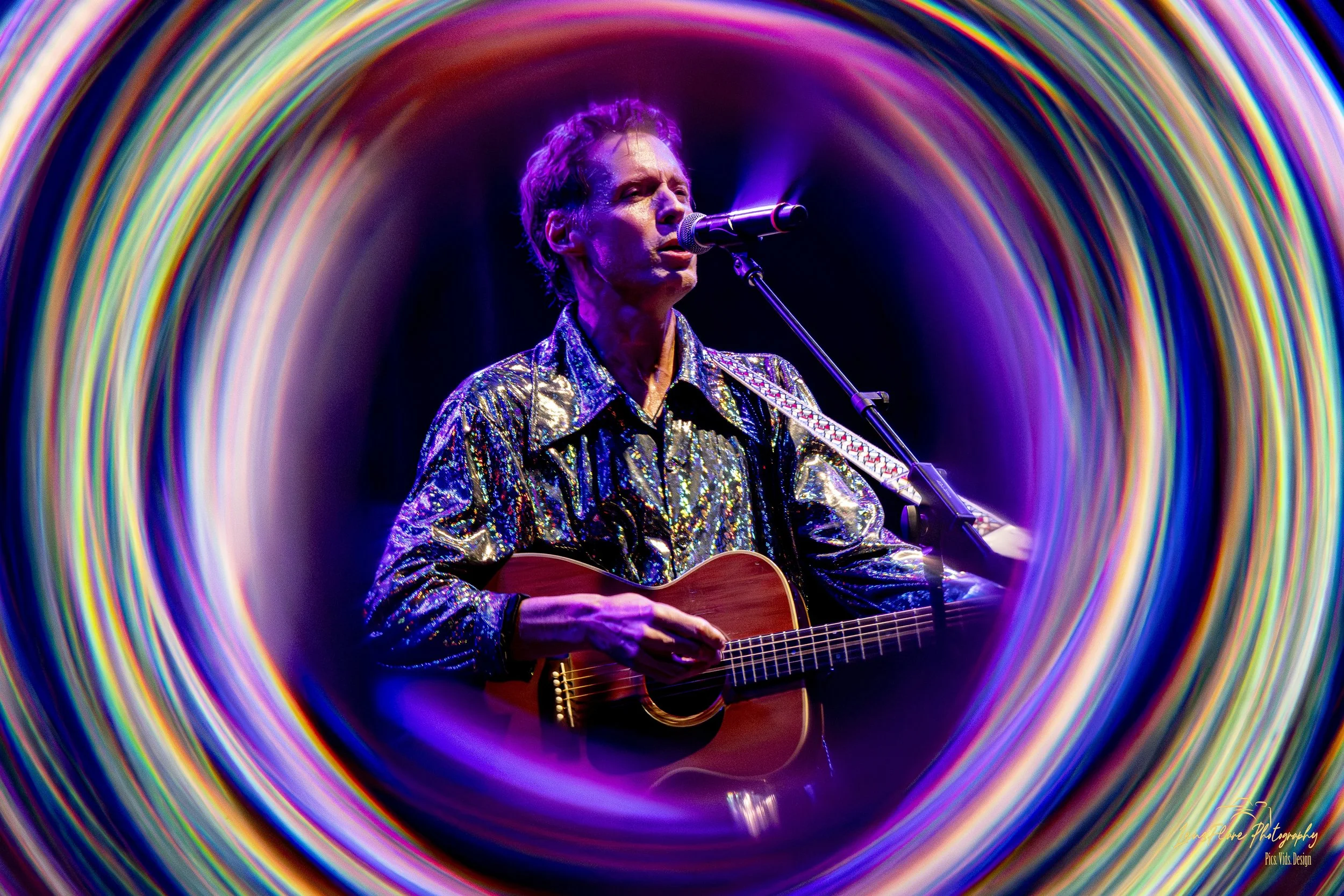 A male musician performing with a guitar, singing into a microphone, wearing a shiny, colorful jacket, framed by concentric rainbow-colored circular lights.