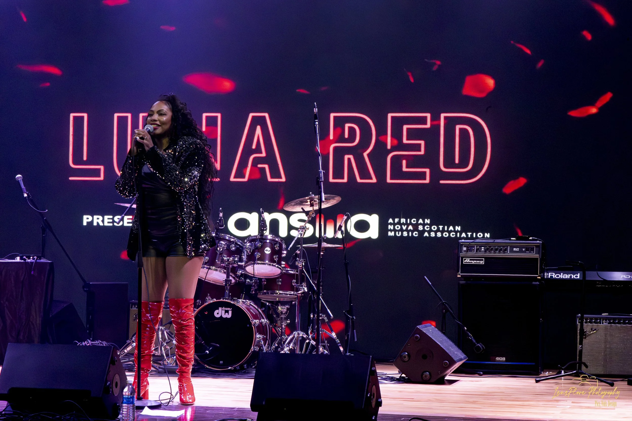 A woman in a sparkly black jacket and red shiny boots holding a microphone on stage with musical instruments and a neon sign in the background.