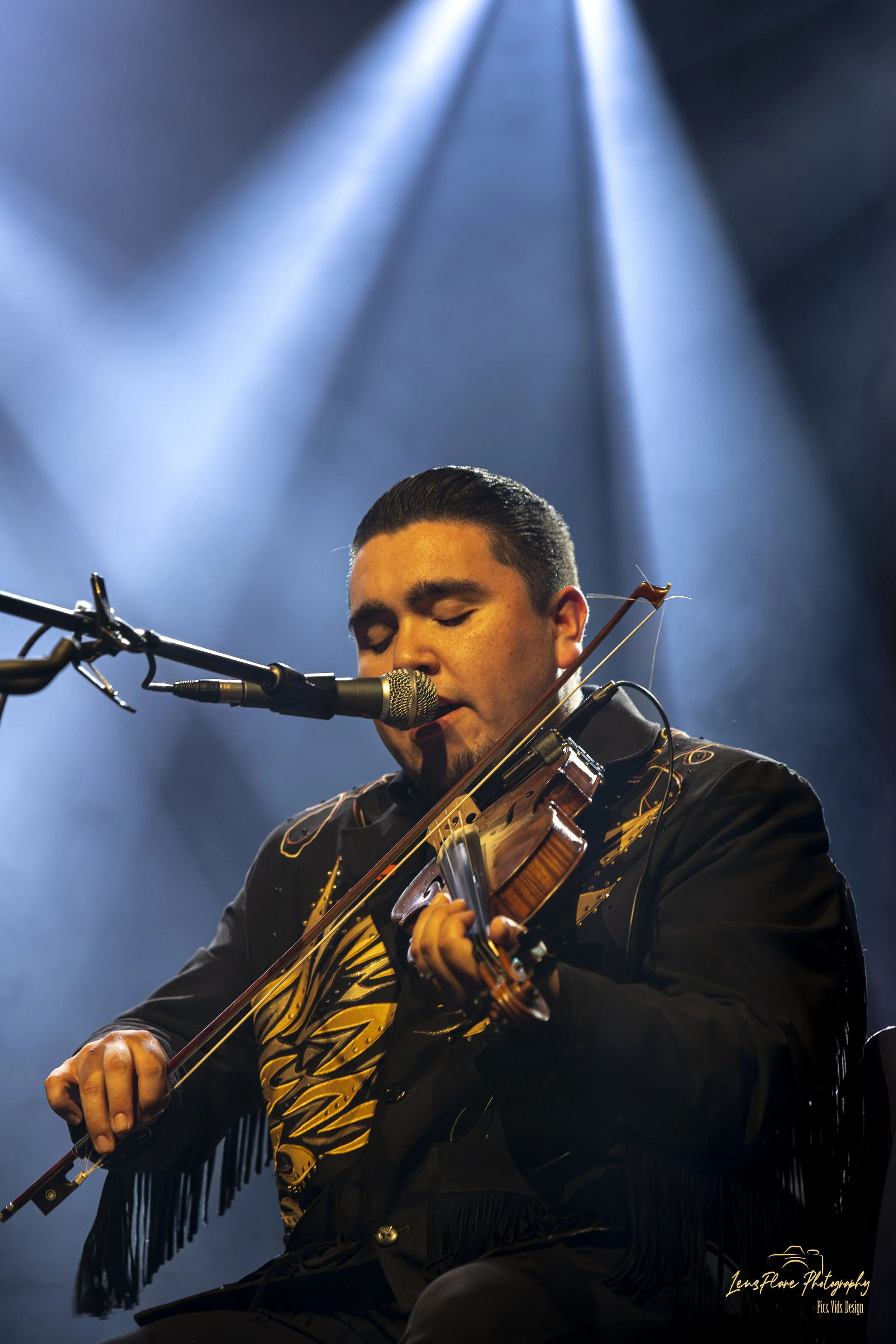 A man playing the violin on stage, illuminated by blue stage lights. He is wearing a black jacket with yellow flame-like designs and black pants, with his eyes closed and focused on his performance.