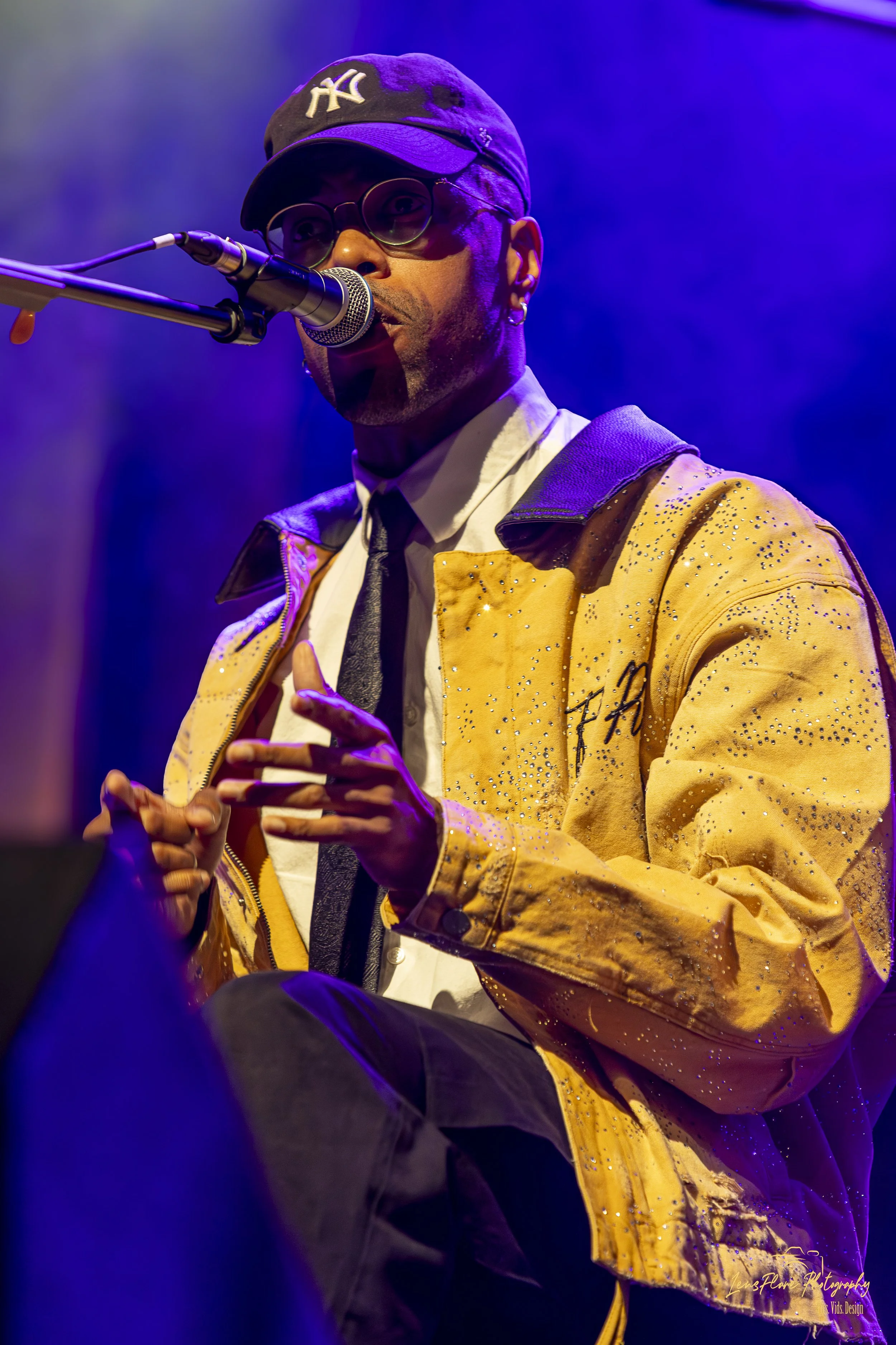 A man performing on stage, wearing a yellow jacket with embellishments, a black cap with white embroidery, glasses, and a white shirt with a black tie, singing into a microphone with purple lighting in the background.