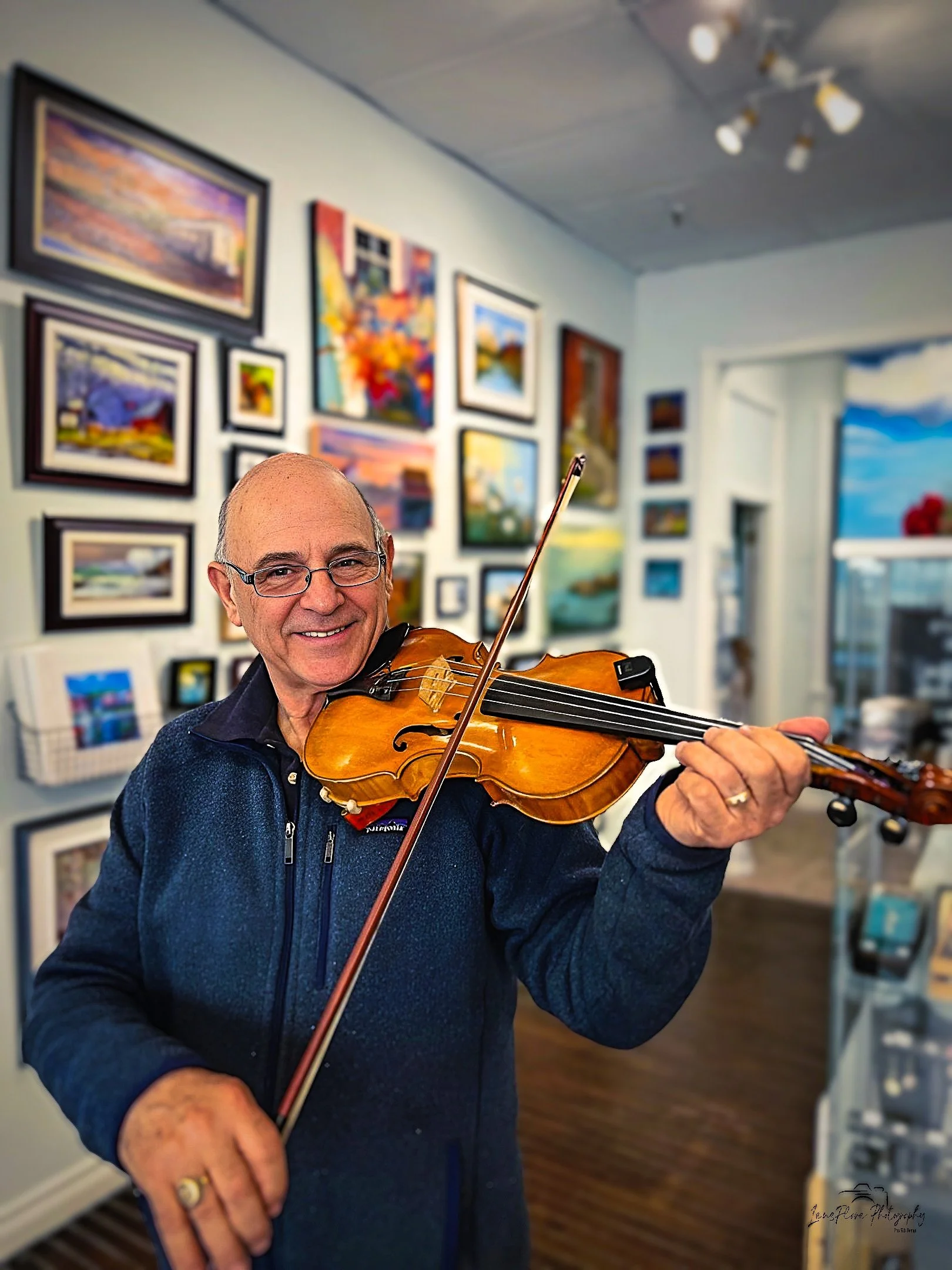 An elderly man with glasses smiling while playing a violin in an art gallery with colorful framed paintings on the wall behind him.