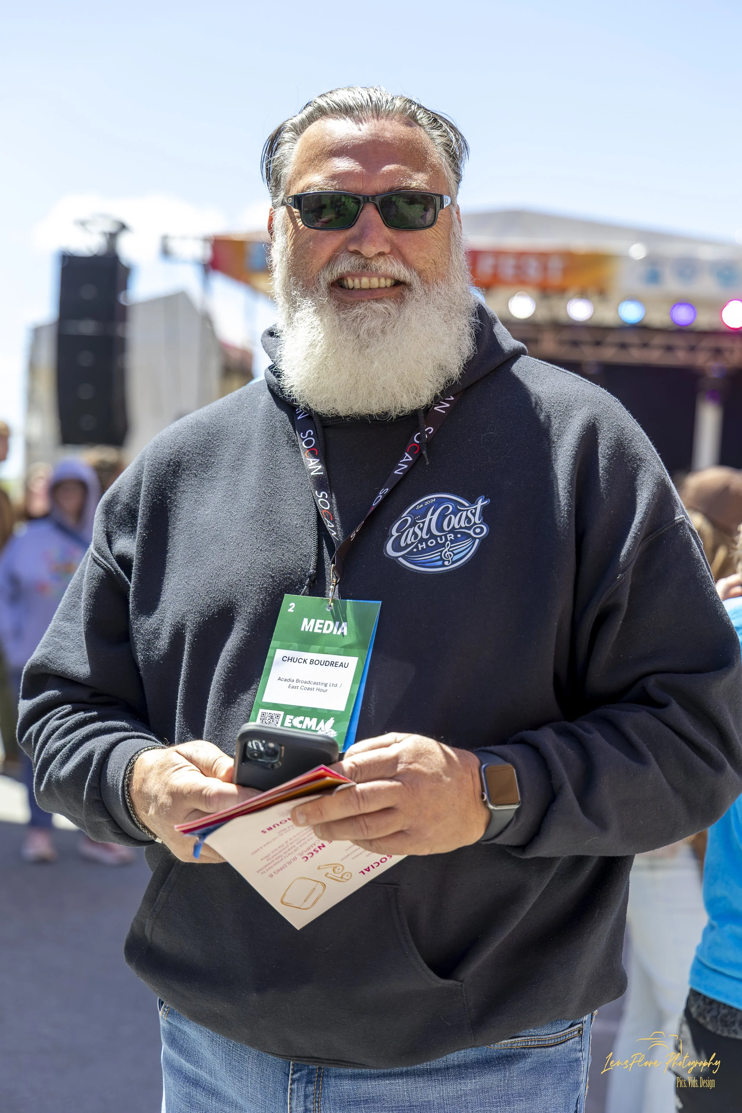 A middle-aged man with a long white beard and sunglasses, wearing a dark jacket with a media pass around his neck, is standing outdoors at a festival or event, holding a smartphone and some printed material, with other people and a stage with lights 