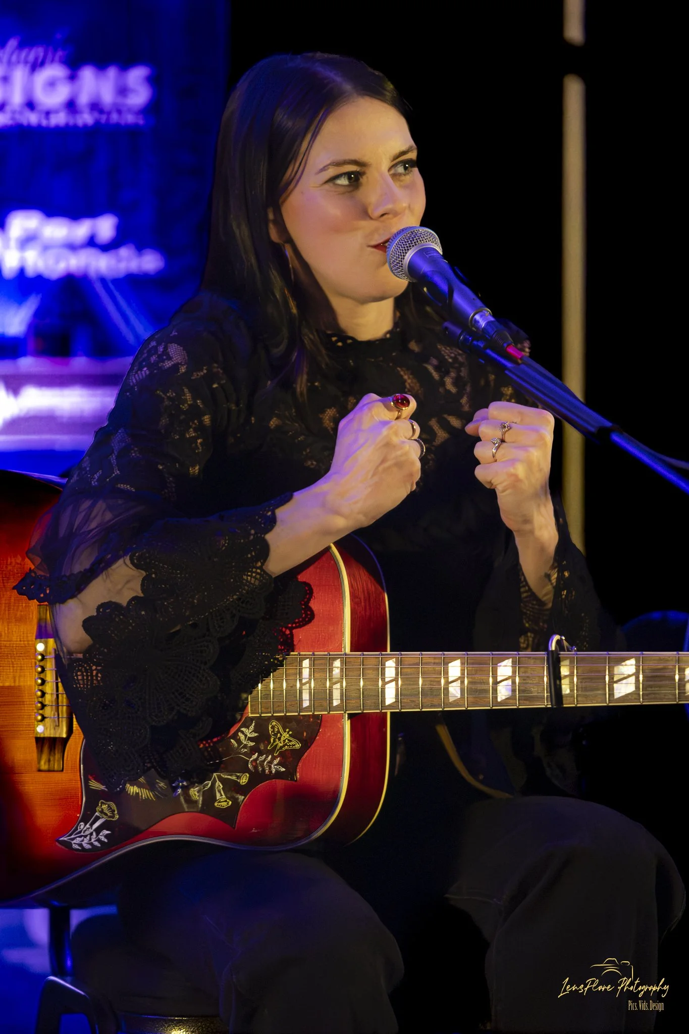 A woman with dark hair wearing a black lace top, sitting on a stage, playing an acoustic guitar with floral stickers, while singing into a microphone with a passionate expression.