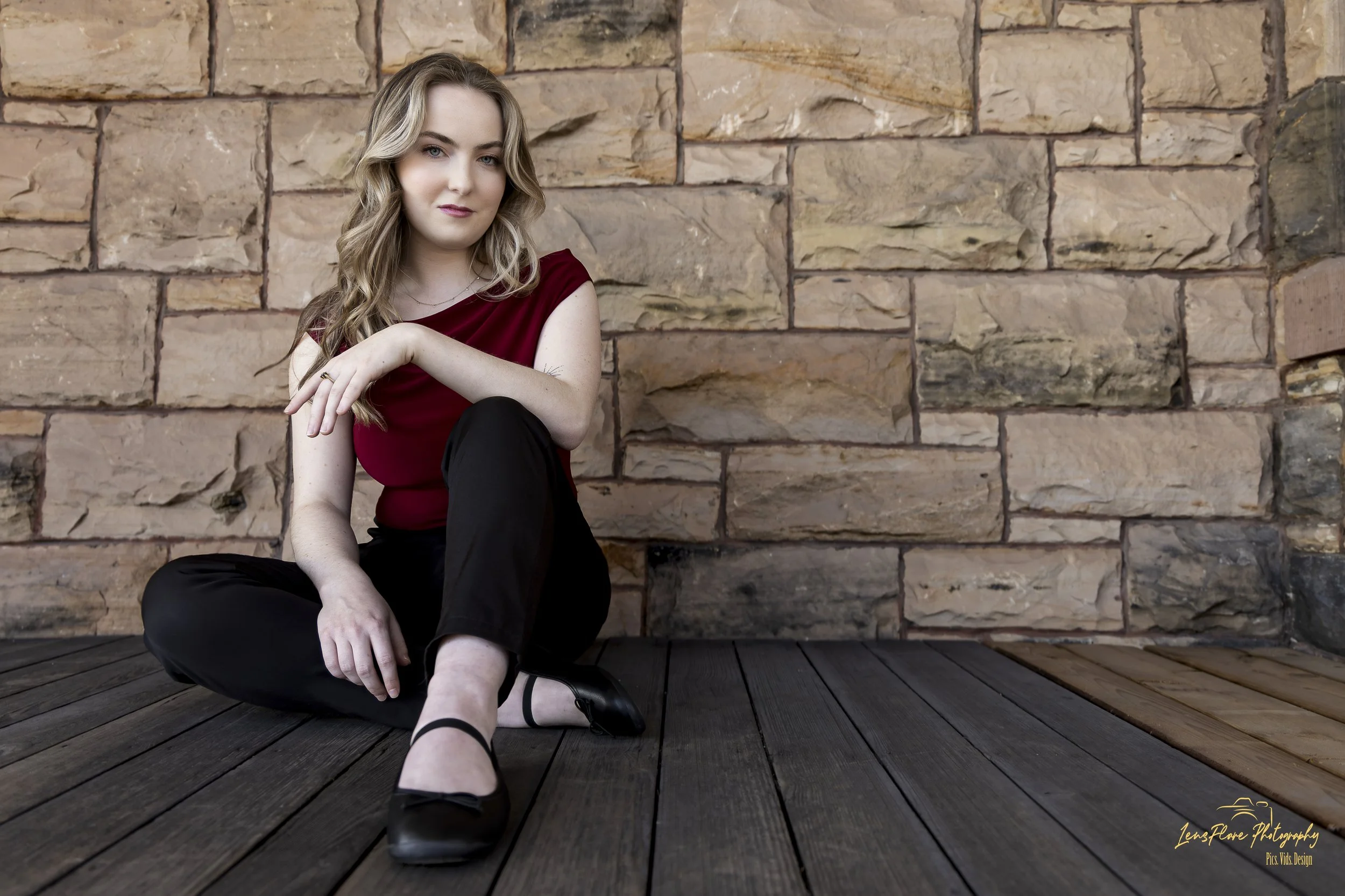 A young woman with long, wavy blonde hair, wearing a burgundy top and black pants, sits on a wooden floor against a stone wall. She has a subtle smile and is looking at the camera.