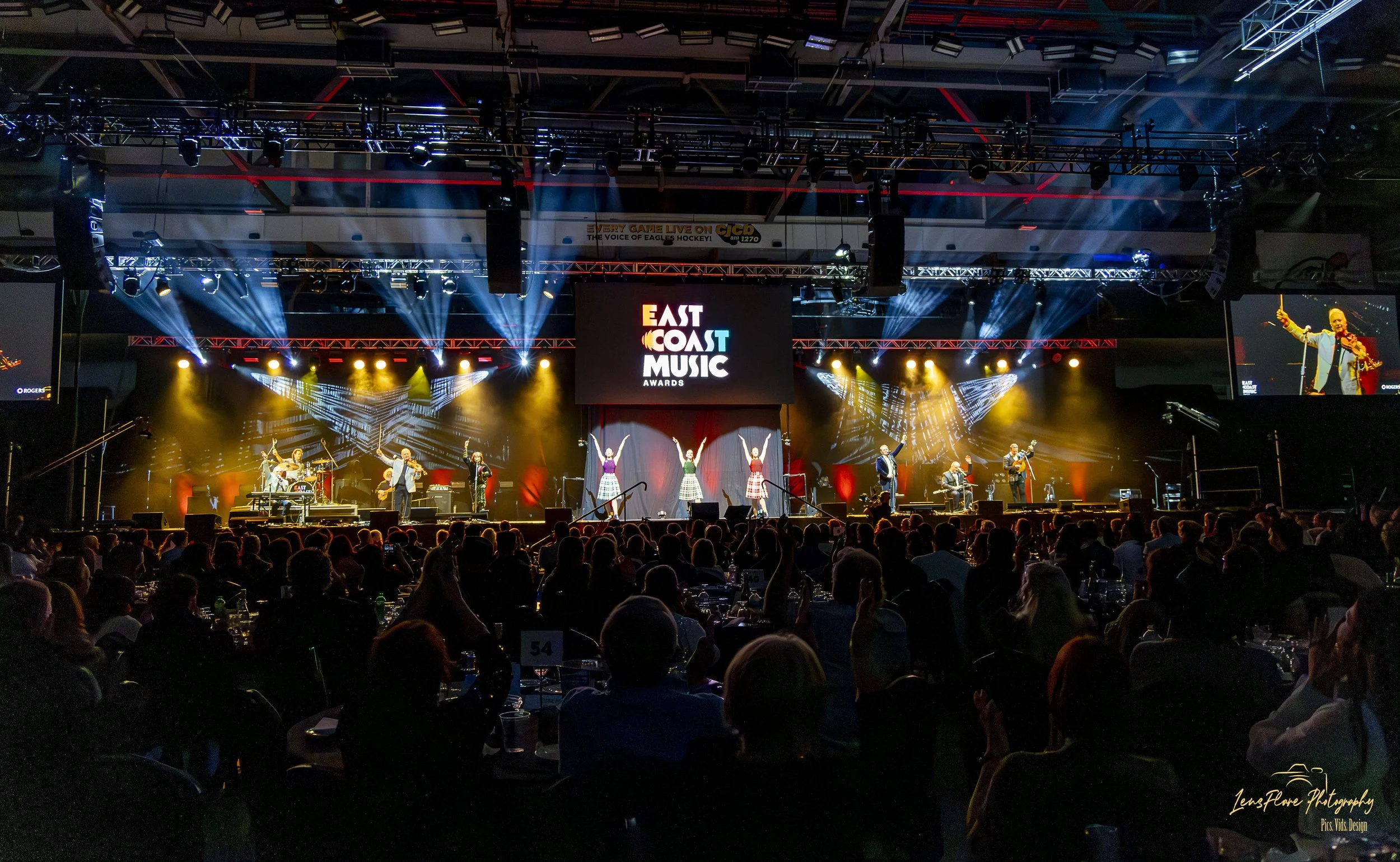 A live music concert at the East Coast Music Awards, with a stage featuring multiple performers, colorful stage lighting, and a large screen displaying the event logo. The audience is seated at tables in the foreground.