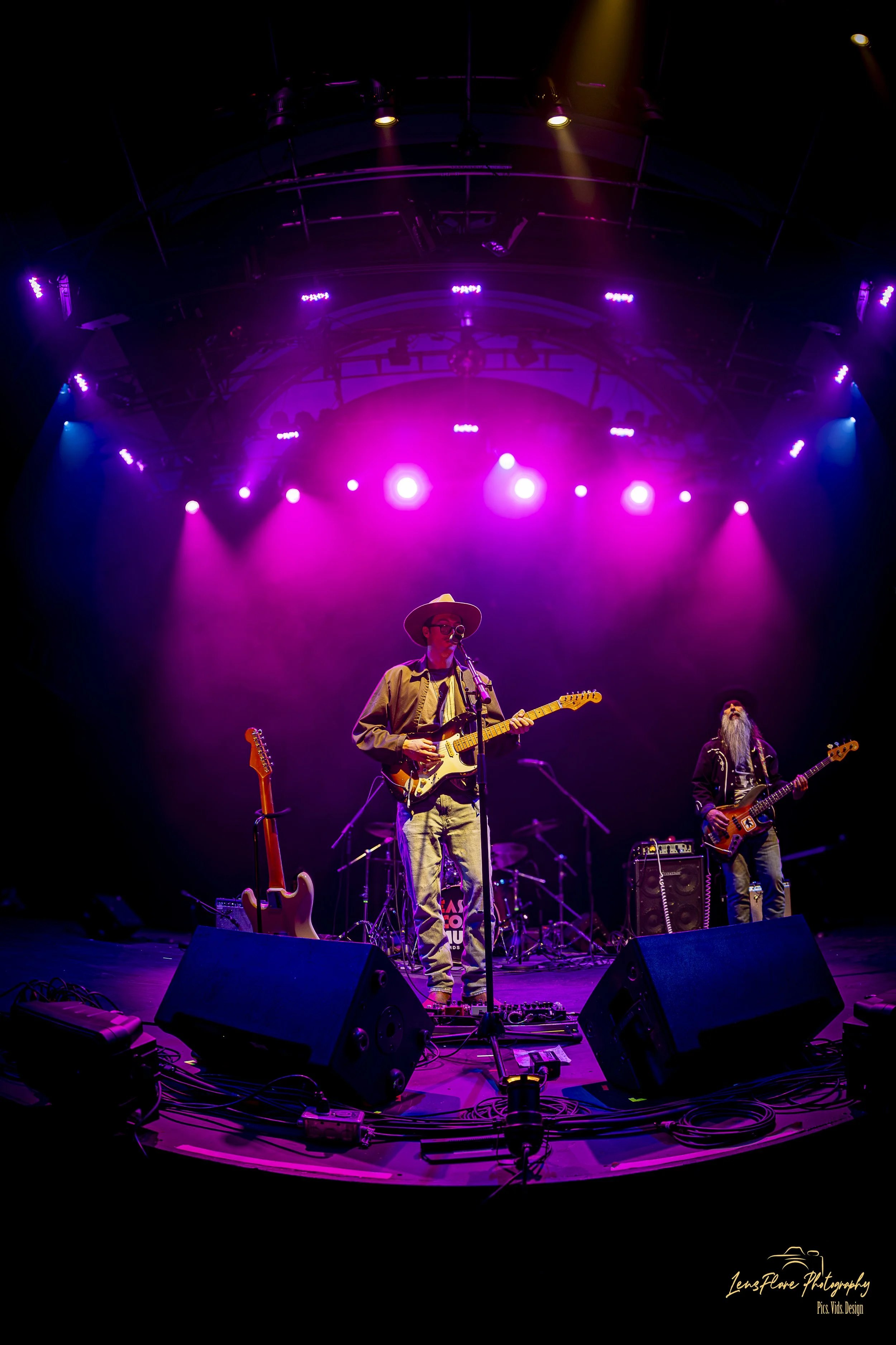 Musicians performing on stage with colorful purple, pink, and blue lighting, featuring two guitarists, one singing into a microphone, and musical equipment visible.
