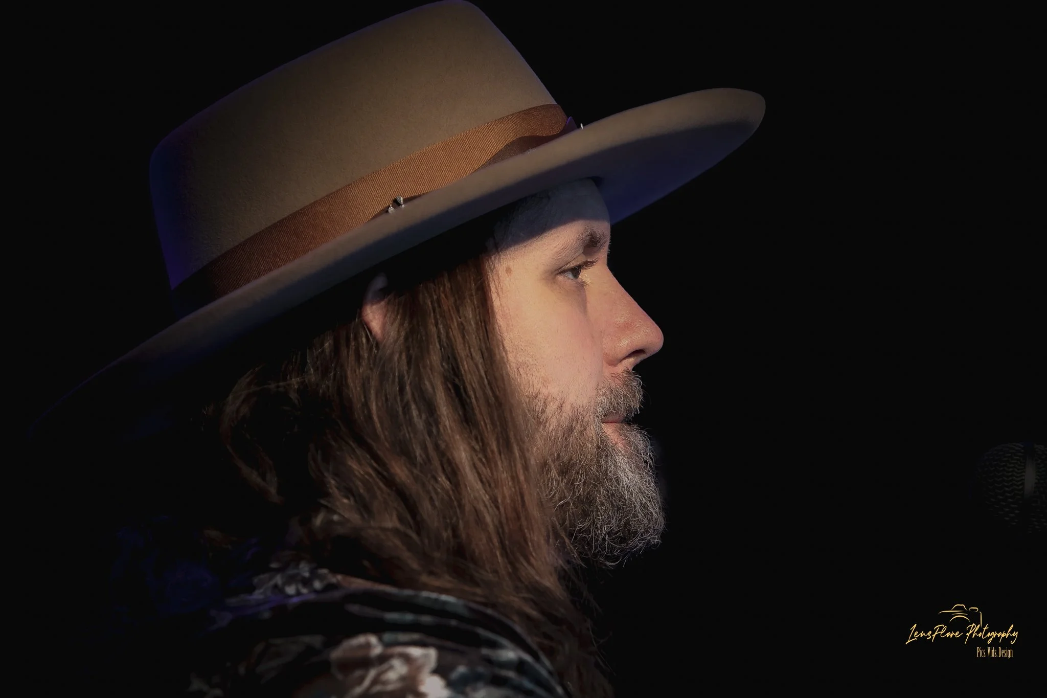 Close-up side profile of a man with long hair and a beard wearing a wide-brimmed beige hat with brown band, against a dark background.