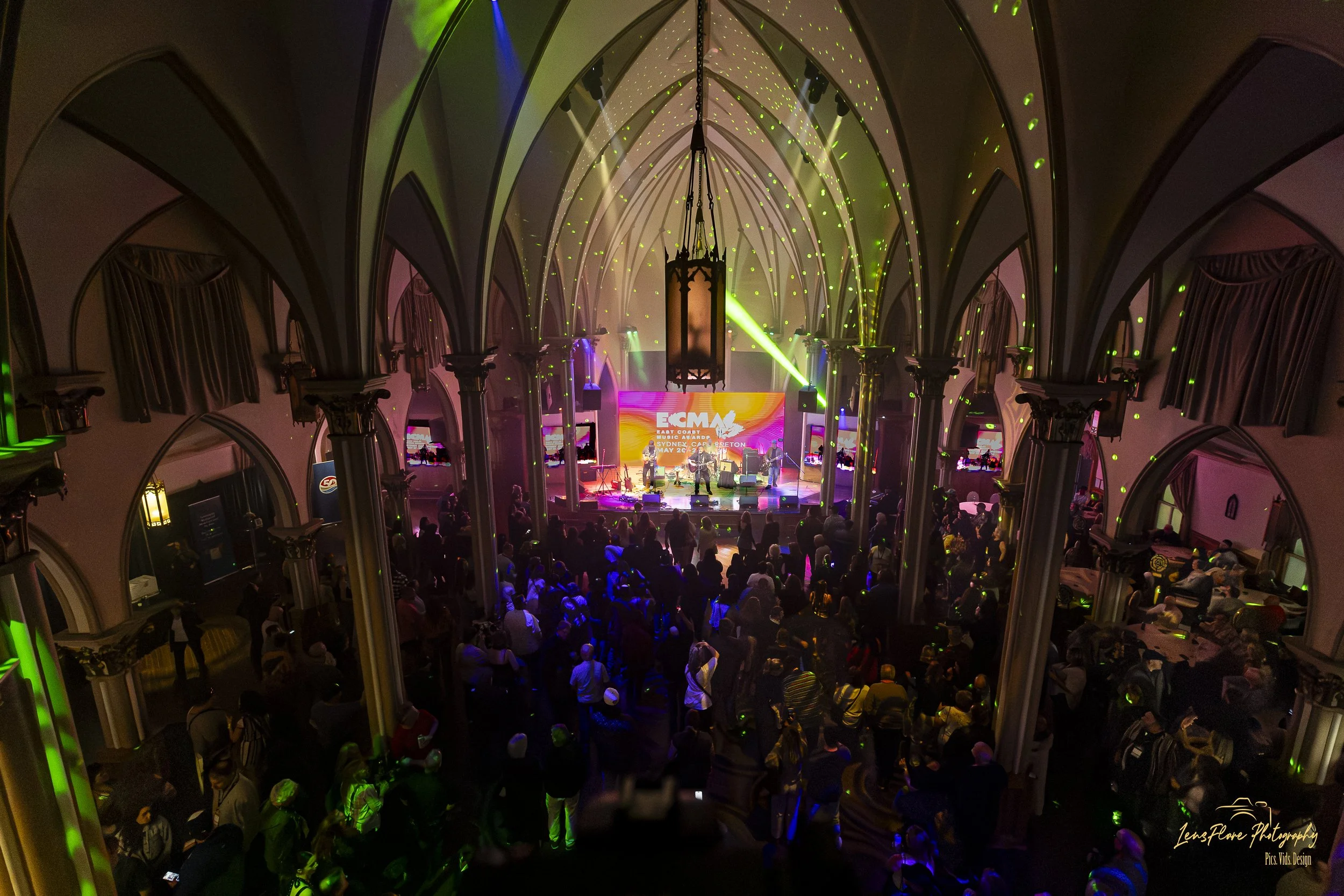 An indoor concert hall with high arched ceilings, columns, and balconies filled with people. A band is performing on stage with colorful lighting and a digital screen displaying the event information.