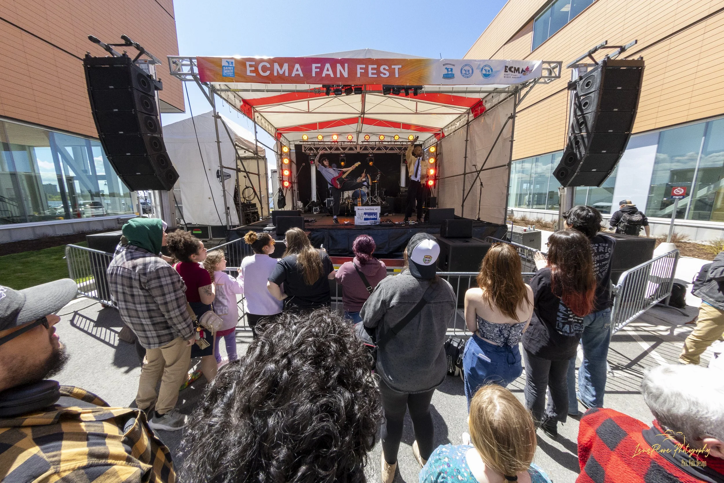 Outdoor concert stage with band playing. Audience gathered in front, surrounded by tall buildings under clear blue sky. Banner reads 'ECMA FAN FEST'.